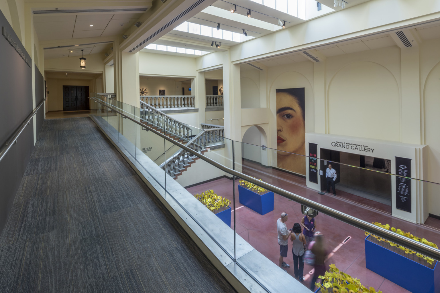 A carpeted walkway overlooks a staircase and an entryway labeled Grand Gallery in cream colored room with skylights