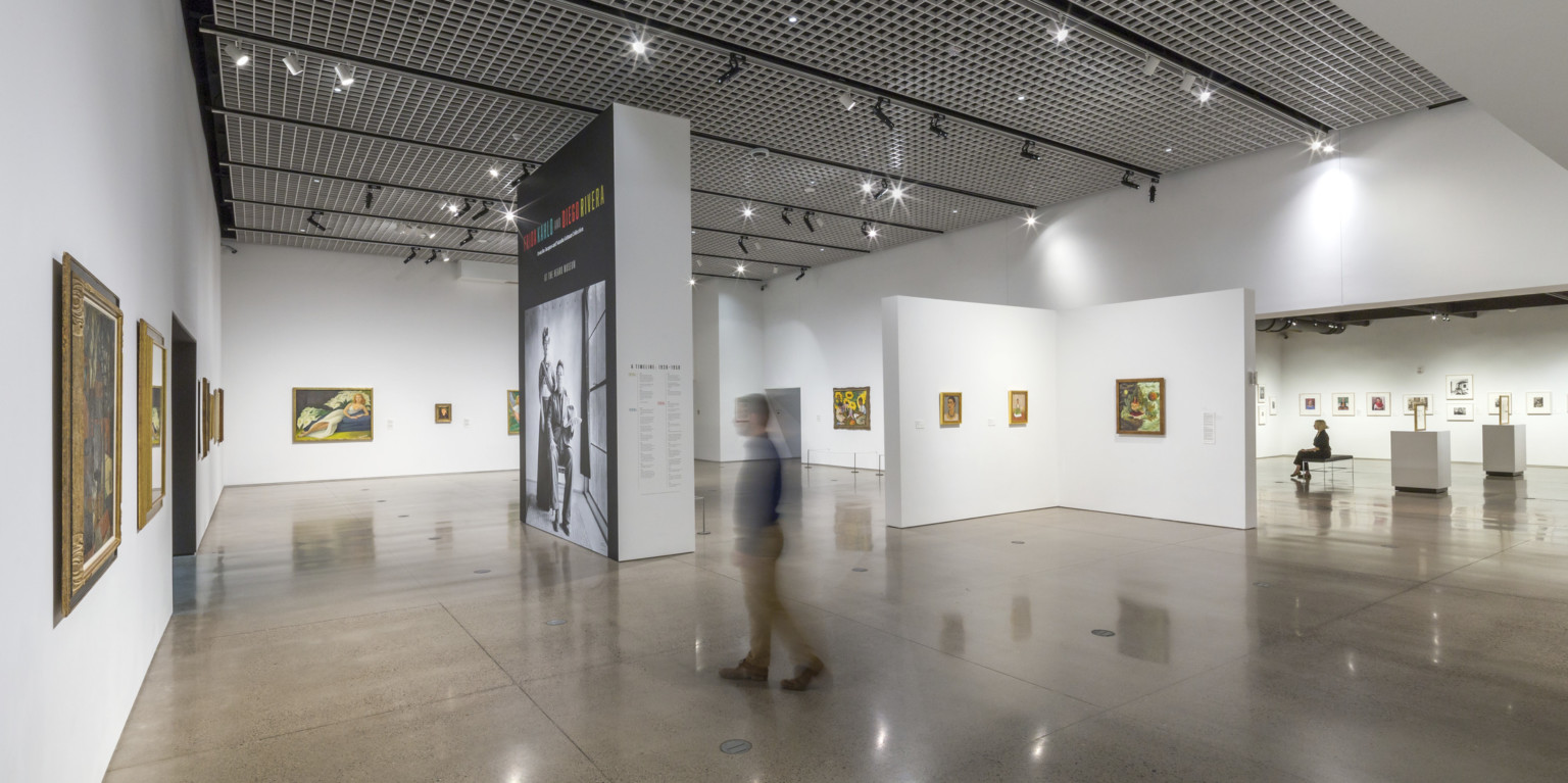 A white gallery in the Heard Museum with paintings displayed on the walls and a view of the adjoining gallery