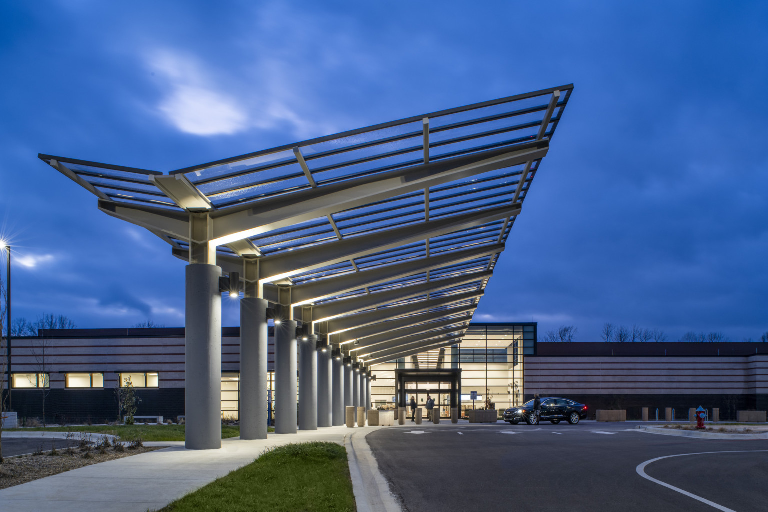 Entry viewed from driveway below angled transparent canopy with metal grid supported by grey pillars with large trusses