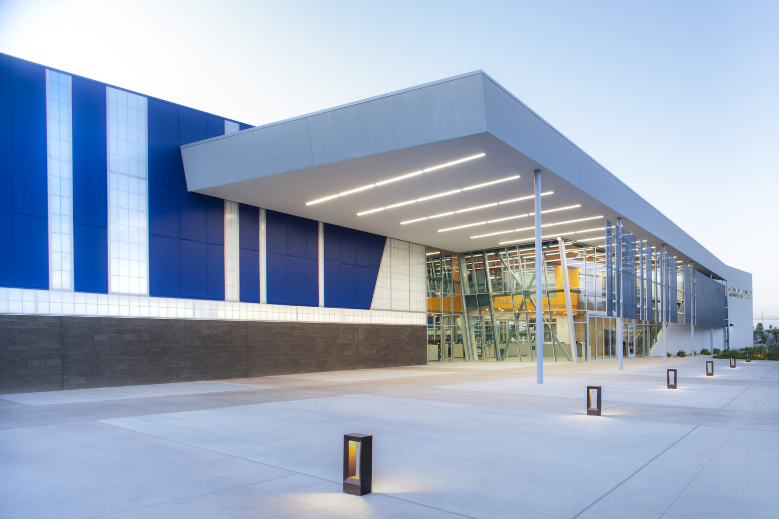 Entry with lit walkway in front of translucent walled building with blue panels and illuminated canopy overhang