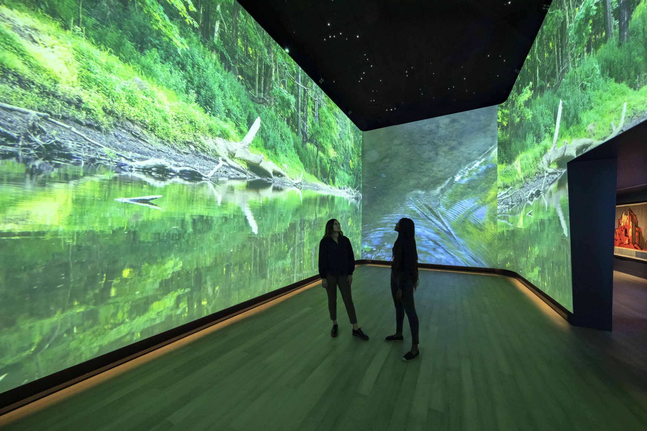 An immersive art exhibit at the Heard Museum with screens on all visible walls of the room showing a stream surrounded by greenery, interactive museum