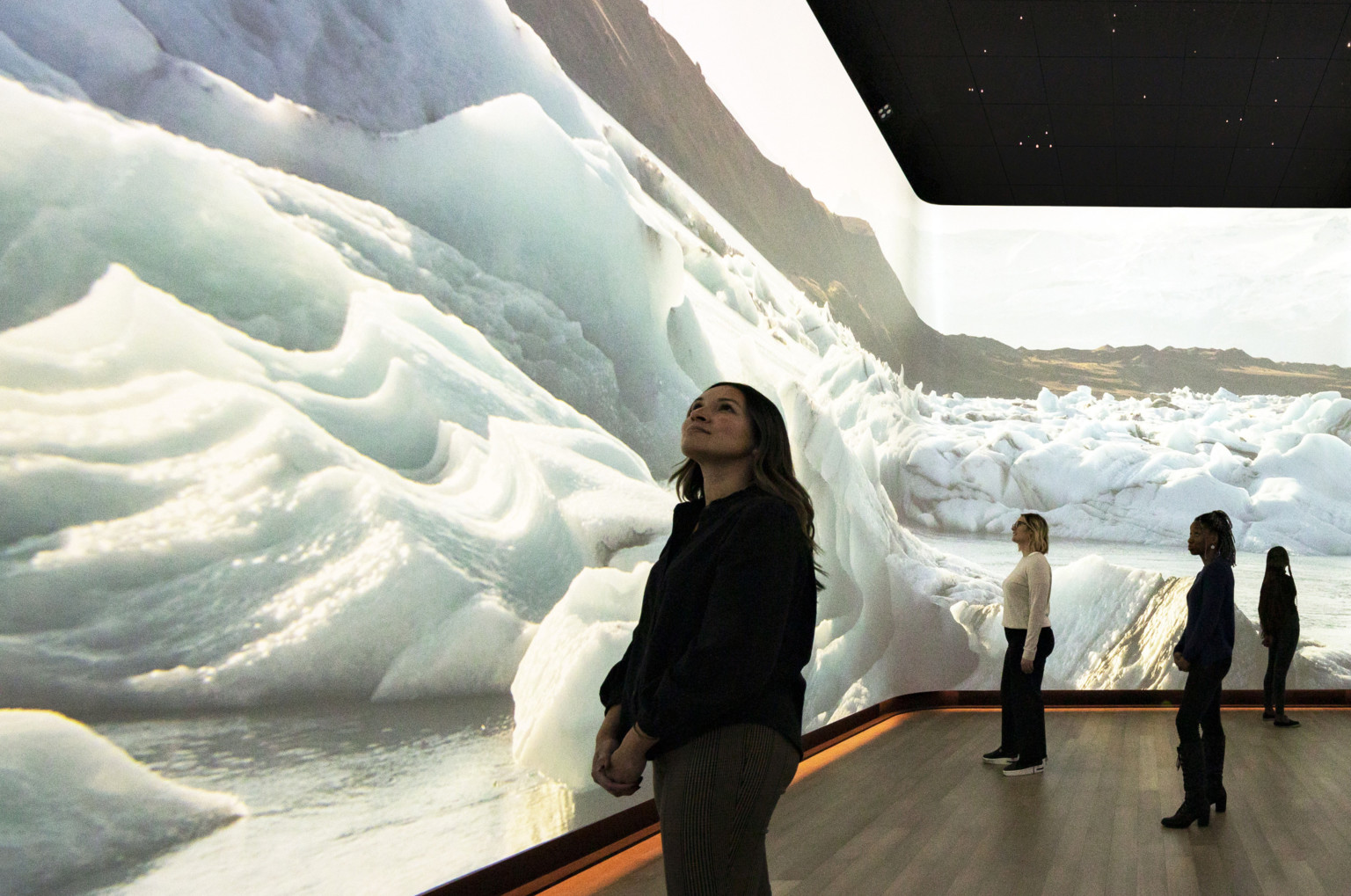 Woman stands in front of immersive 360 degree video art display of snow dunes and ice, interactive museum