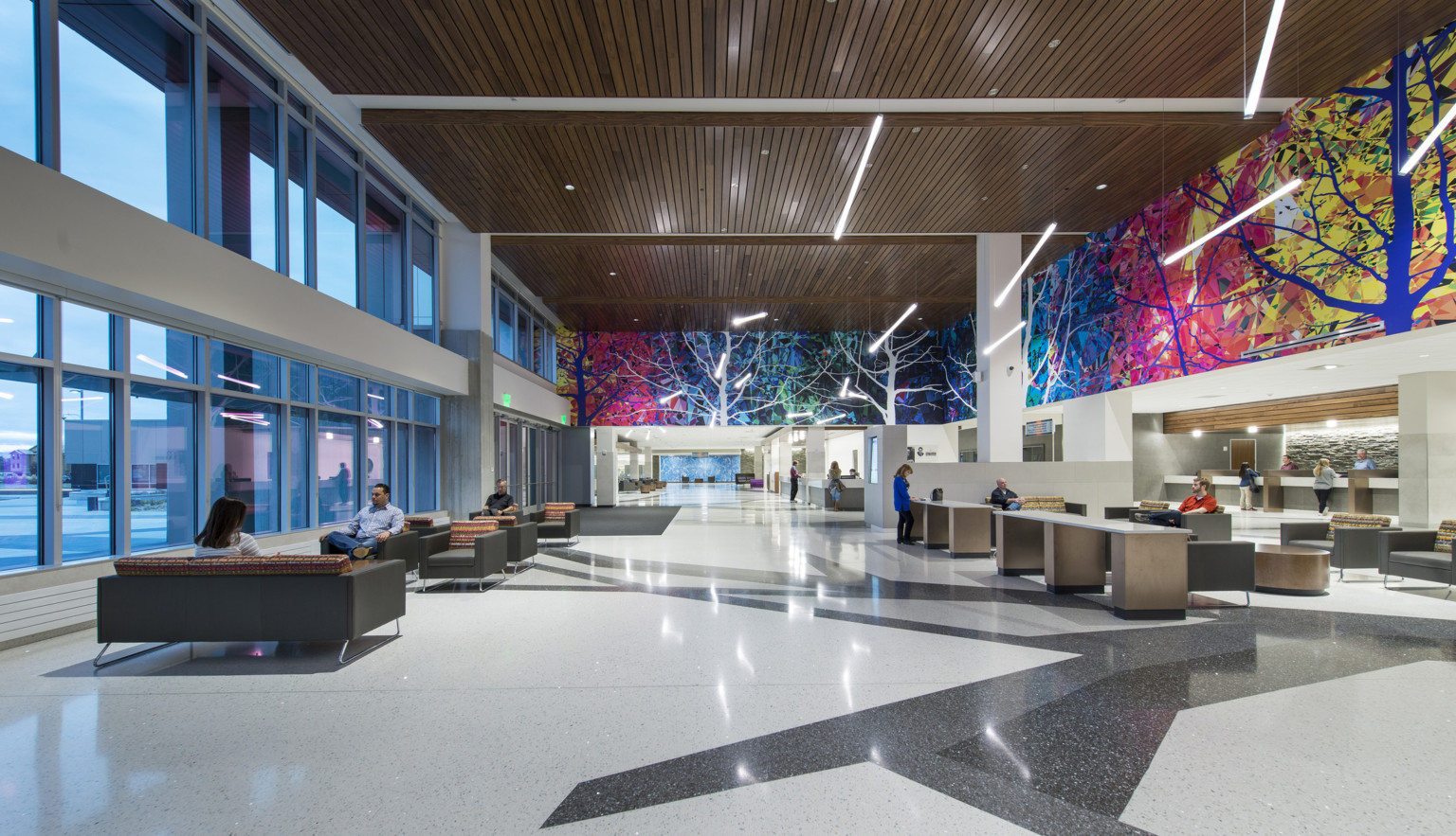 Interior lobby with colorful mural, seating space, desks, and wood detailed ceiling. Tall wall of windows lines the left wall