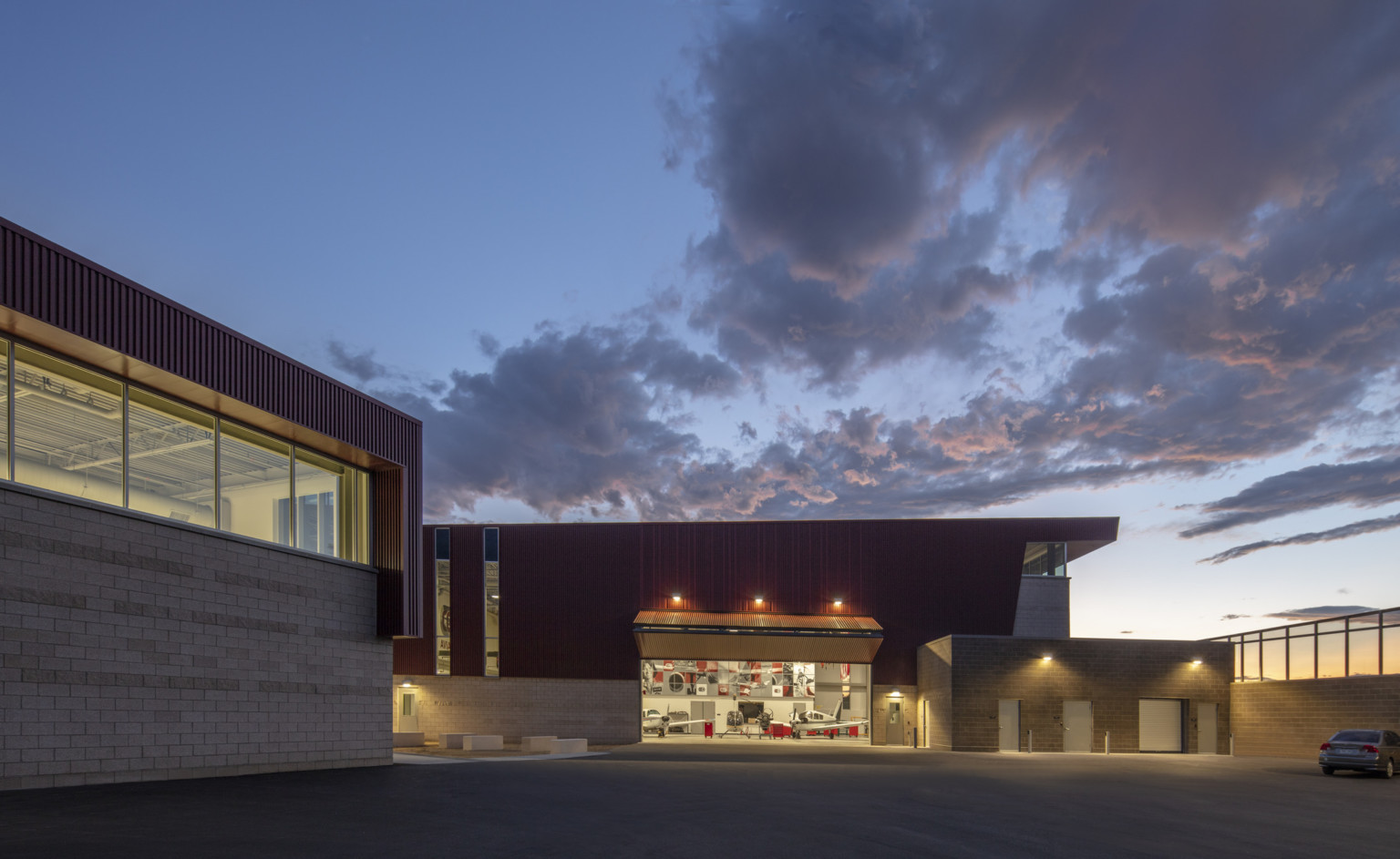 Stone building with textured red wrap facade illuminated from within in evening. Garage door opened at center to airplanes