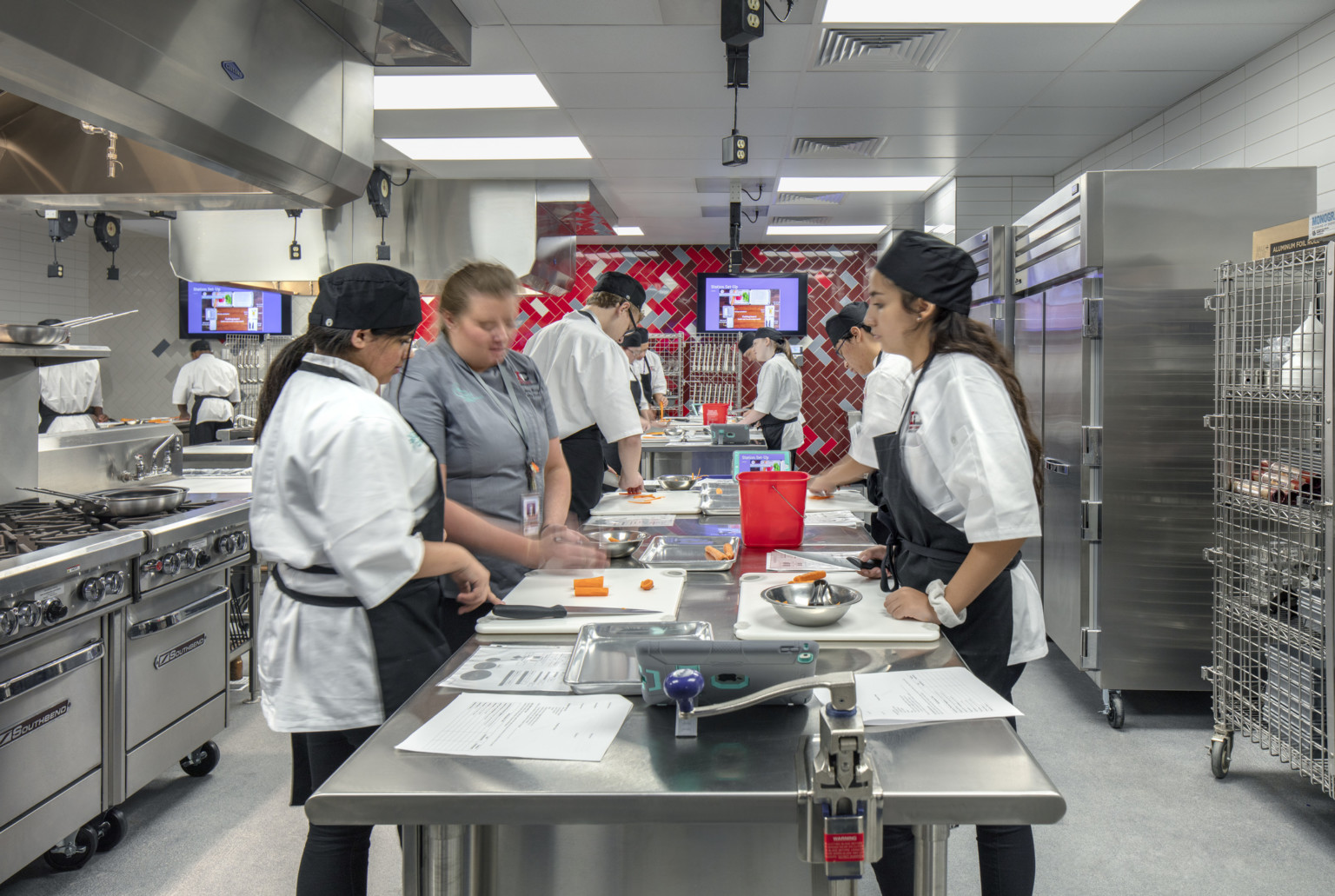 Culinary kitchen classroom with stainless steel island and red tile back wall. Stoves on left wall, refrigerators to right