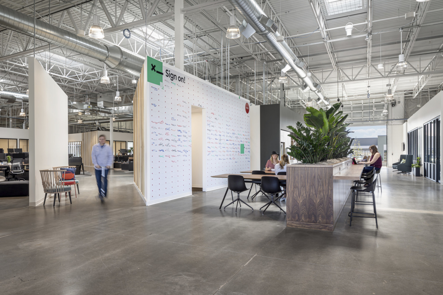 Tables in front of wood half wall with plants on top and counter attached. White board wall to left. Open garage door window