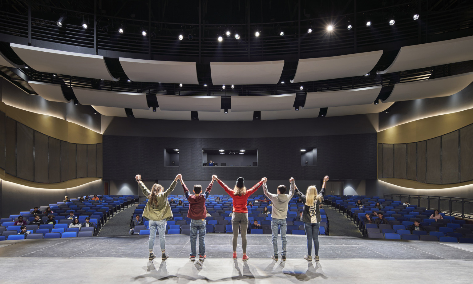 Students stand on stage facing audience. Curved acoustic panels above. Wood details arch along side walls with lights behind Students stand on stage facing audience. Curved acoustic panels above. Wood details arch along side walls with lights behind