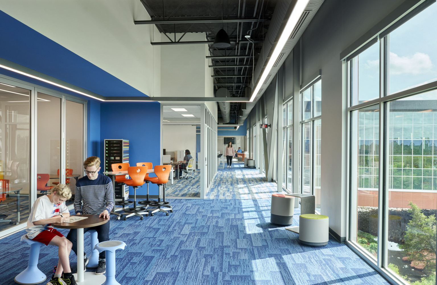 Blue carpeted hall with large windows on right wall. Left, recessed blue wall accent along glass wall looking into classes