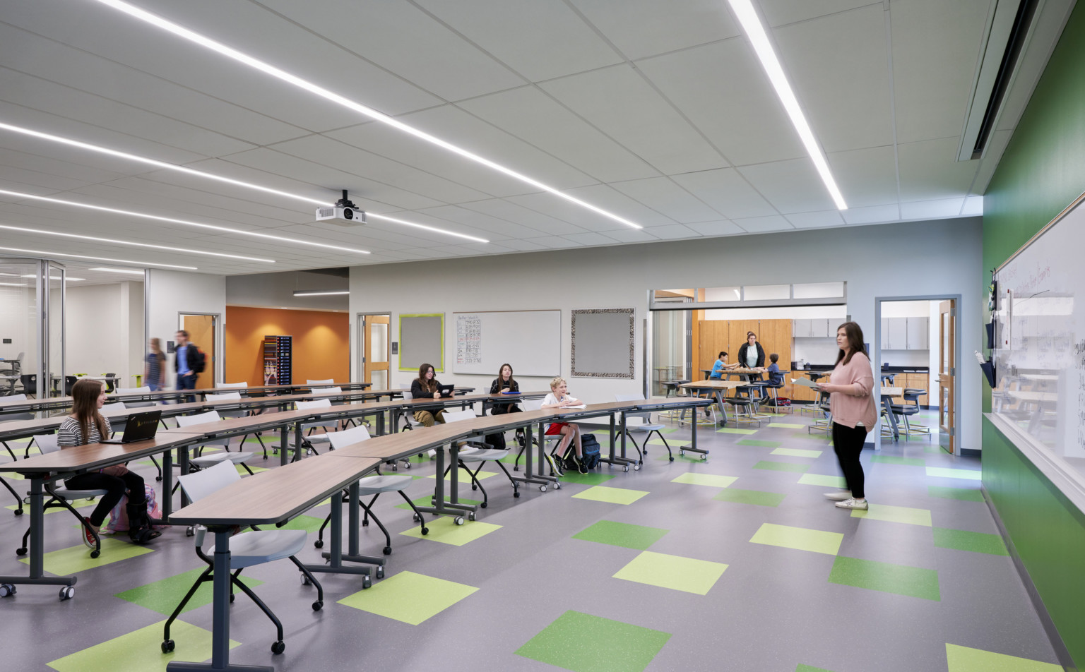 Classroom with green accent wall and floor tiles. Orange walls in hallway through flexible walls. Rolling chairs and long tables