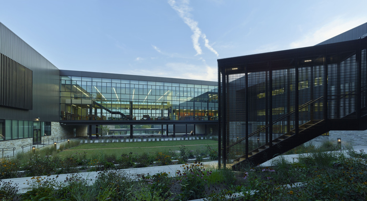 Partially enclosed black stairwell in front of double height raised glass walled hallway. Between is a courtyard with paths Partially enclosed black stairwell in front of double height raised glass walled hallway. Between is a courtyard with paths