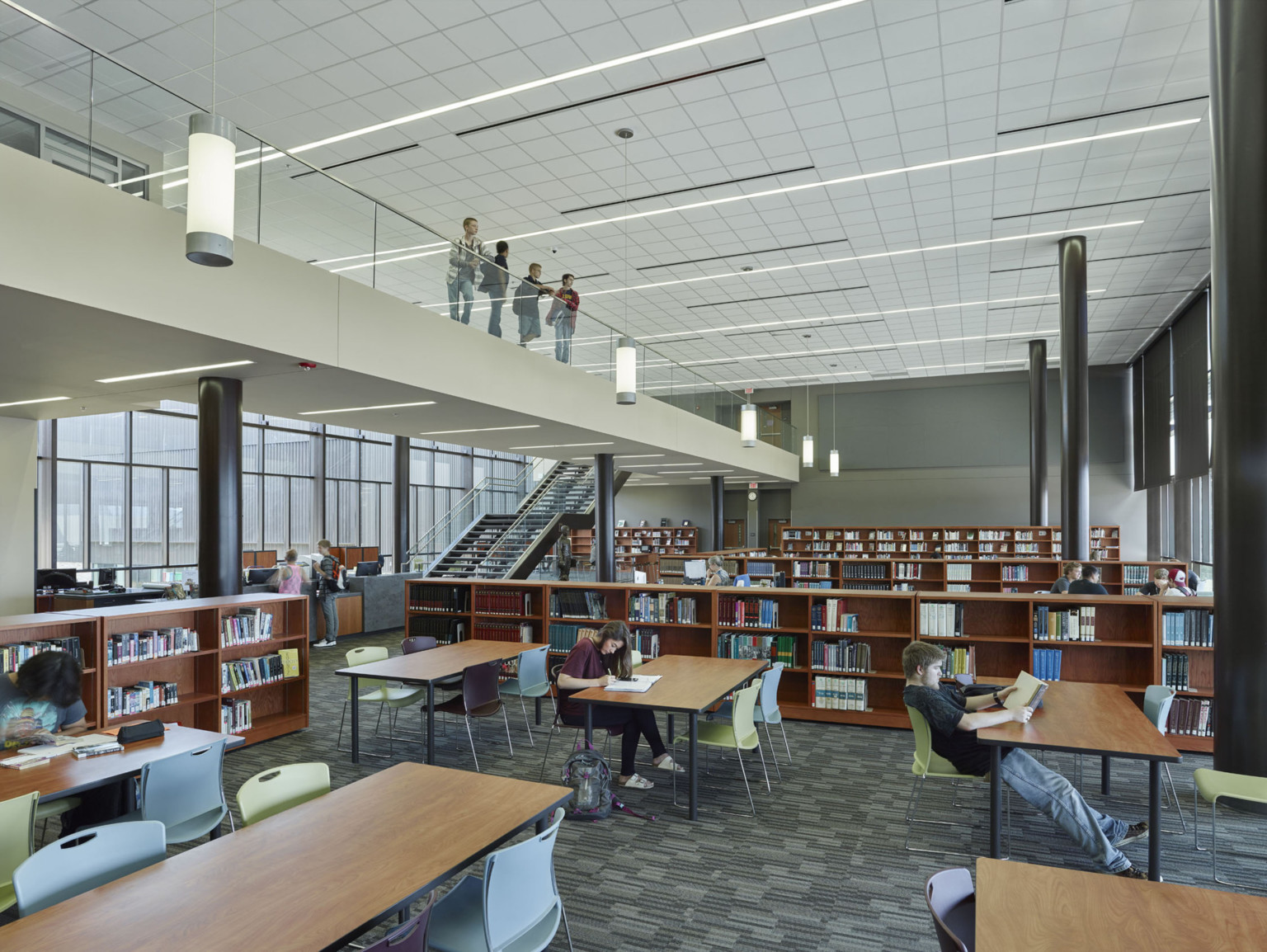 Study area of library surrounded by half height bookshelves. Black columns in double height room with 2nd floor walkway left