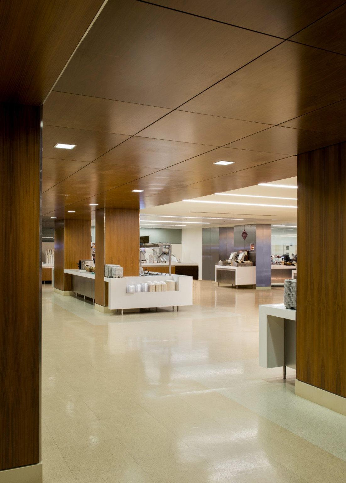 Lobby with cream tiled floor below large wood pillars and ceiling with square recessed lights. White tables and counters.