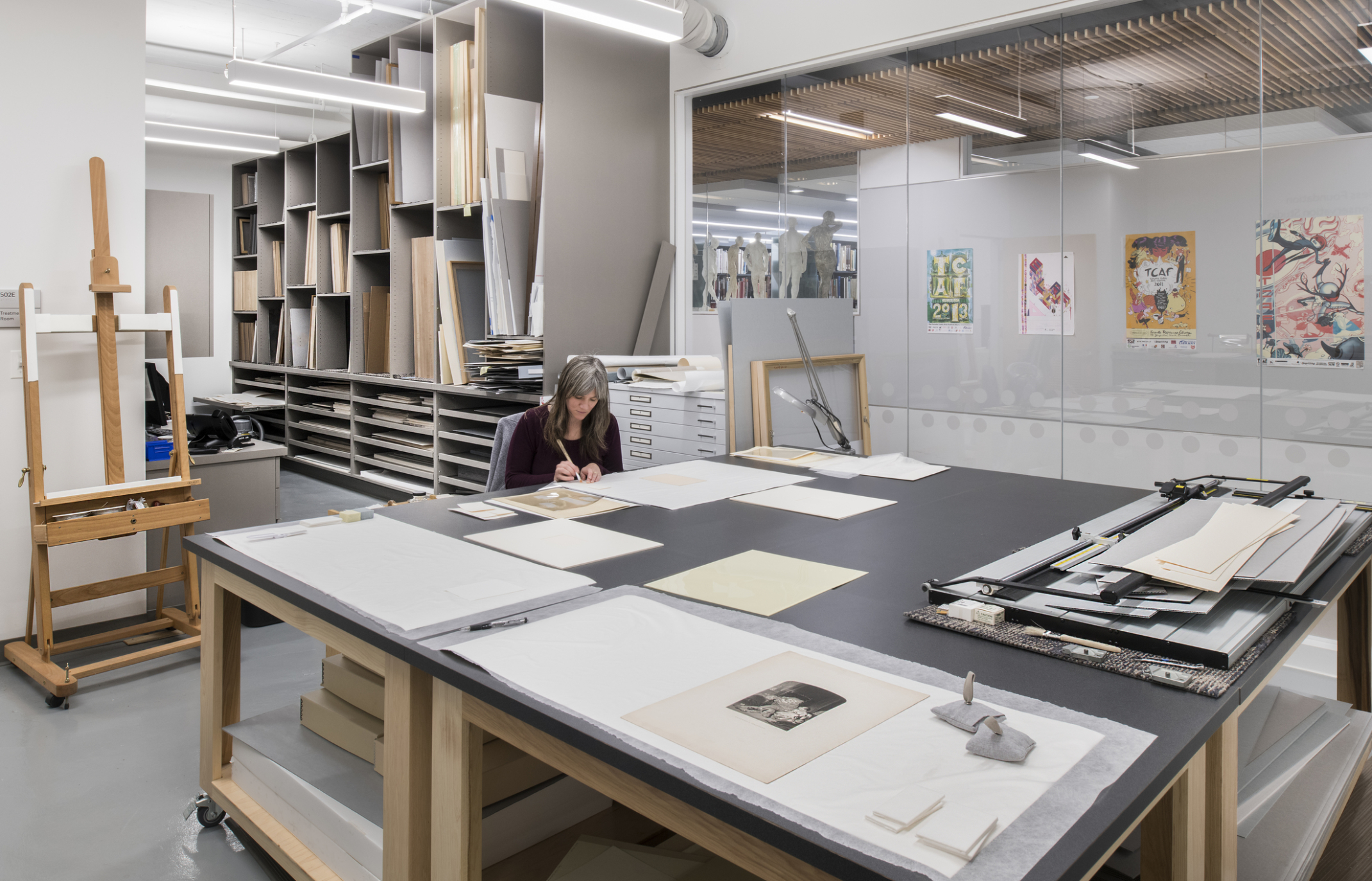 woman sitting at a large work table with artwork, pencil drawings, and sketches laid in front of her. Framed artwork seen through glass wall in the background.