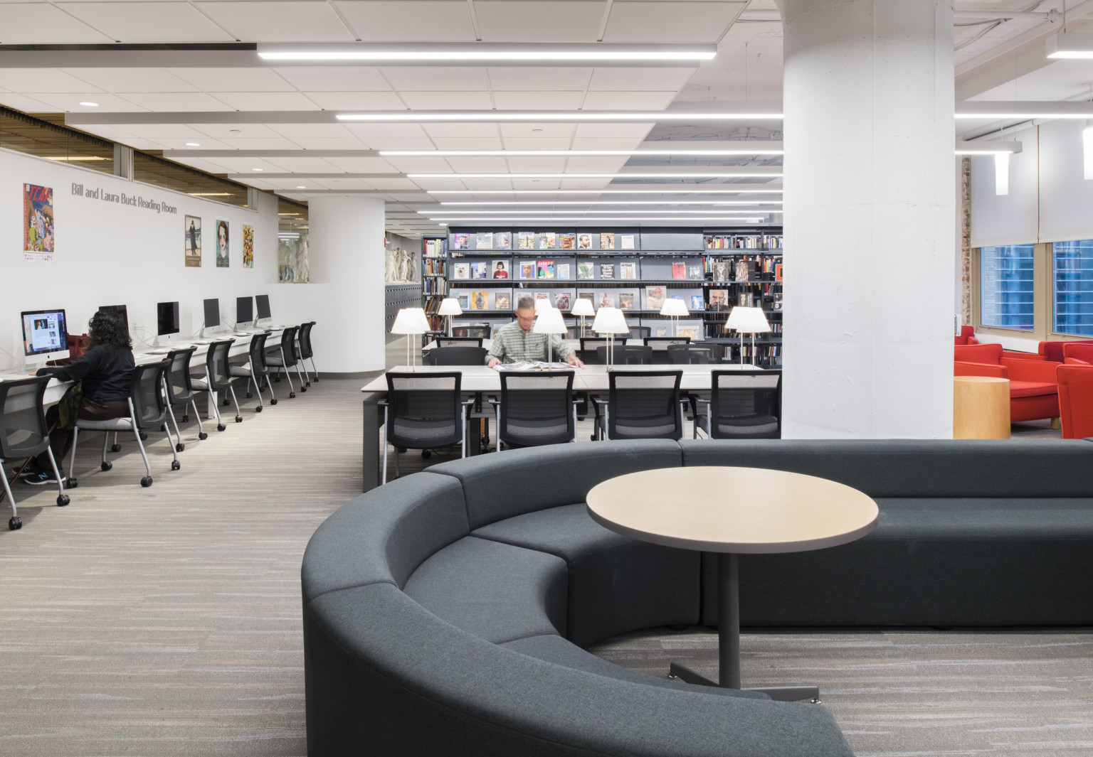 Library with round couch in forefront, tables with reading lamps, shelves of books in back, and lined with table of computers