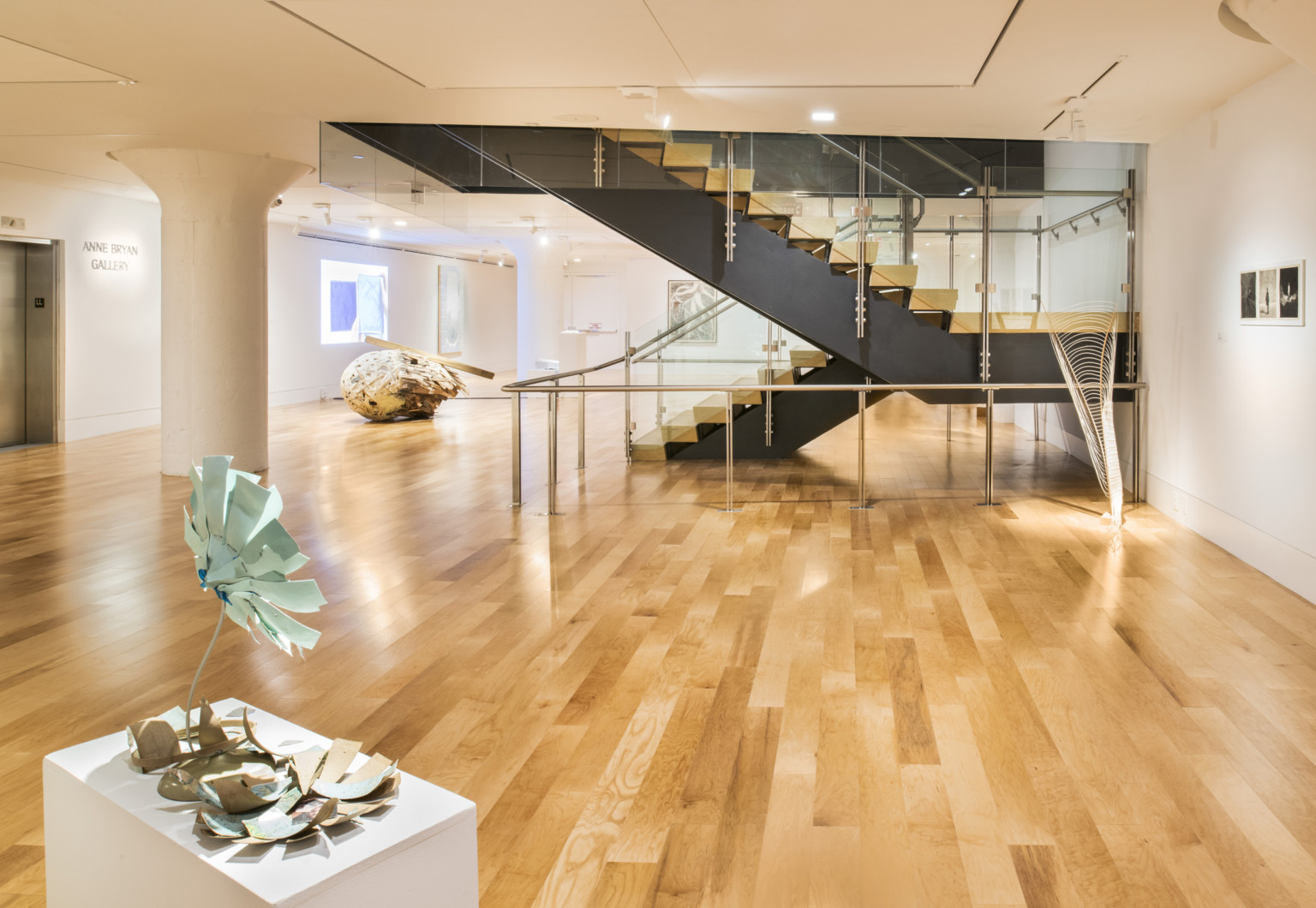 White art gallery with light color hardwood flooring and stairs with black base leading upstairs. Art is on white pedestal