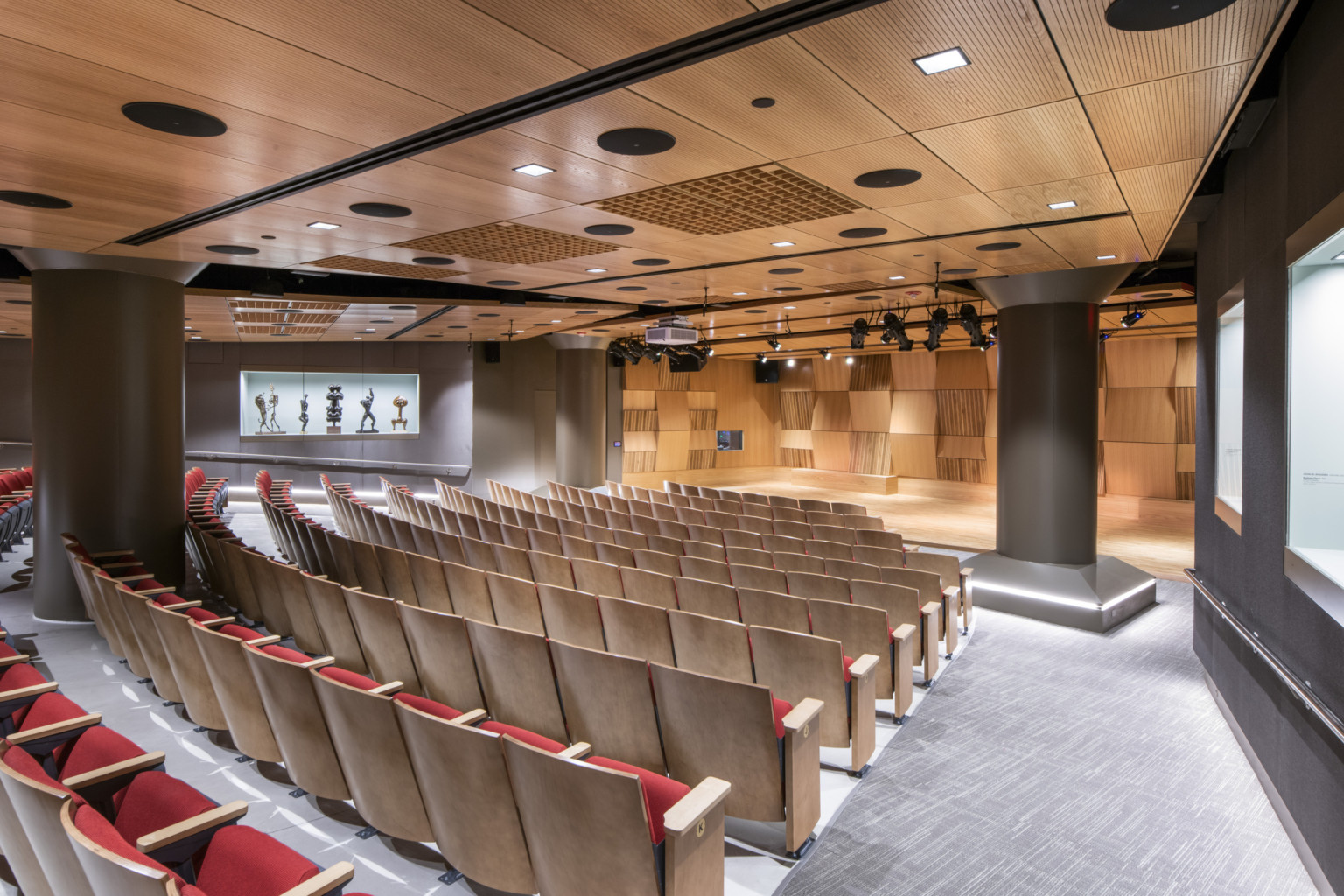 Rows of empty red and wood seats face stage with textural wood walls, lit pockets for art on grey walls near audience seats