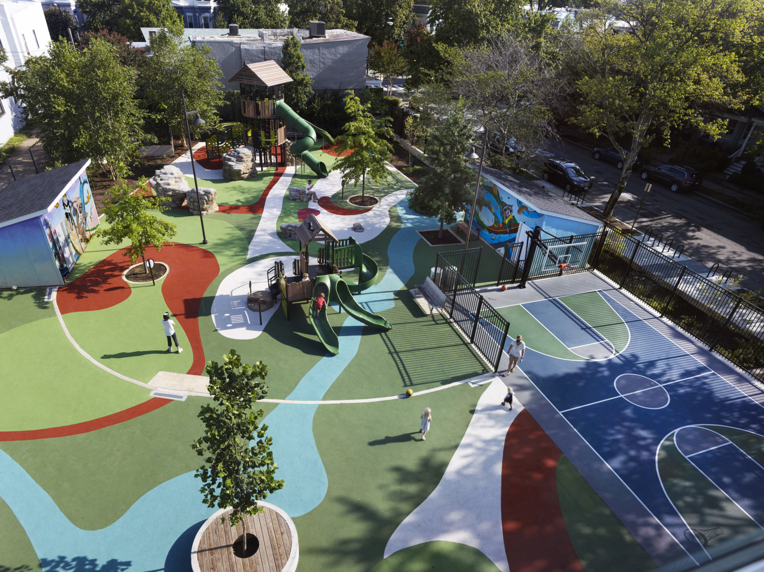 Aerial view of playground with trees, multicolor ground, murals. Blue and green partially fenced in basketball court, right
