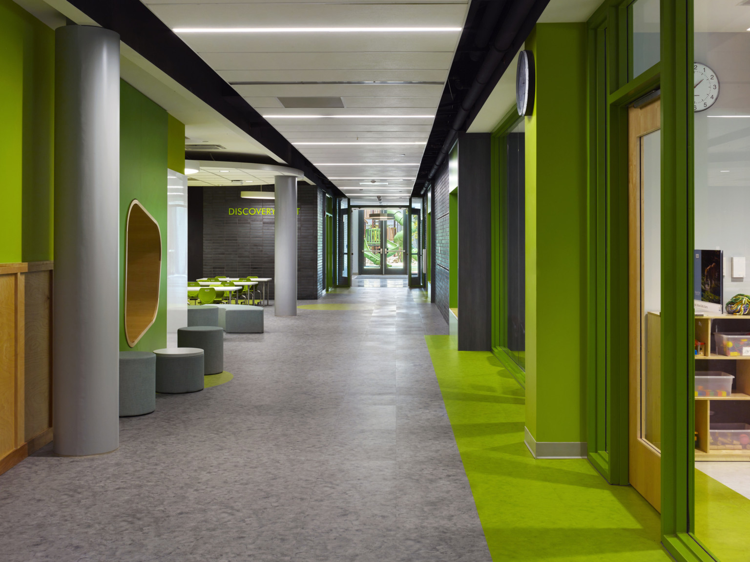 Green hallway with wood accents and transparent classroom walls. Grey stools around organic wood panel wall cutout to left