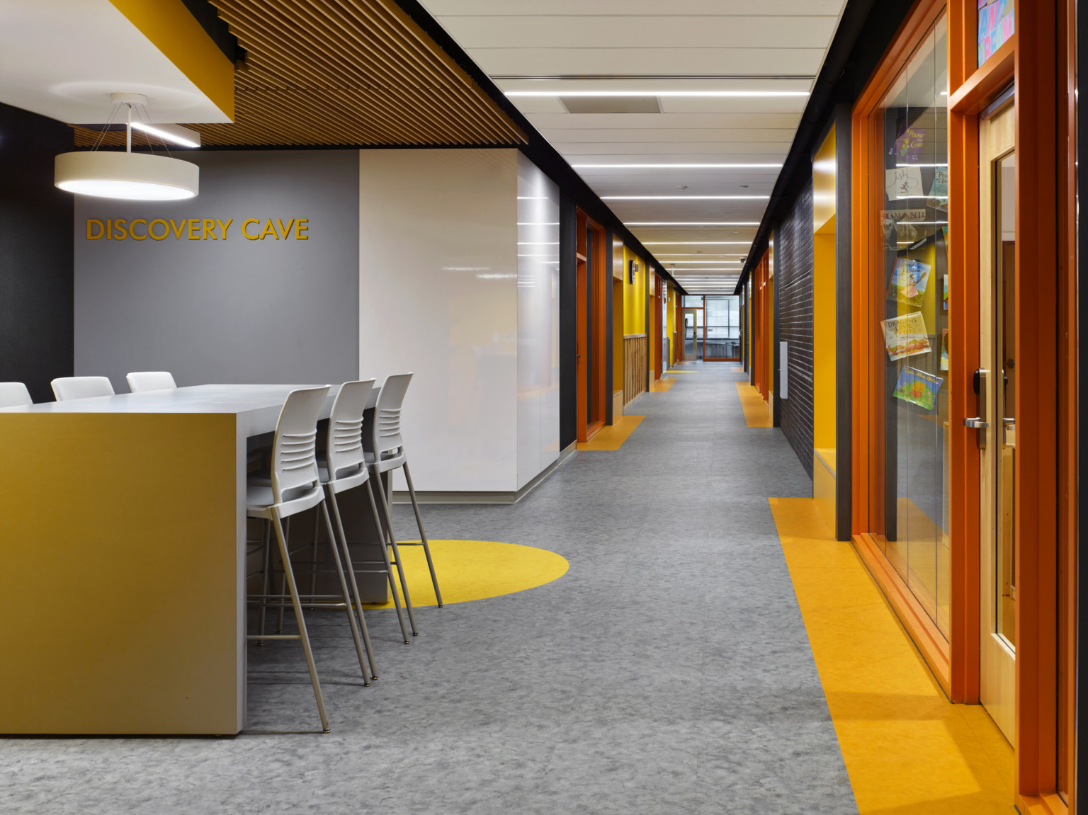 Open room labeled Discovery Cave off hallway. White island table with stools. Wood slat ceiling and yellow accents