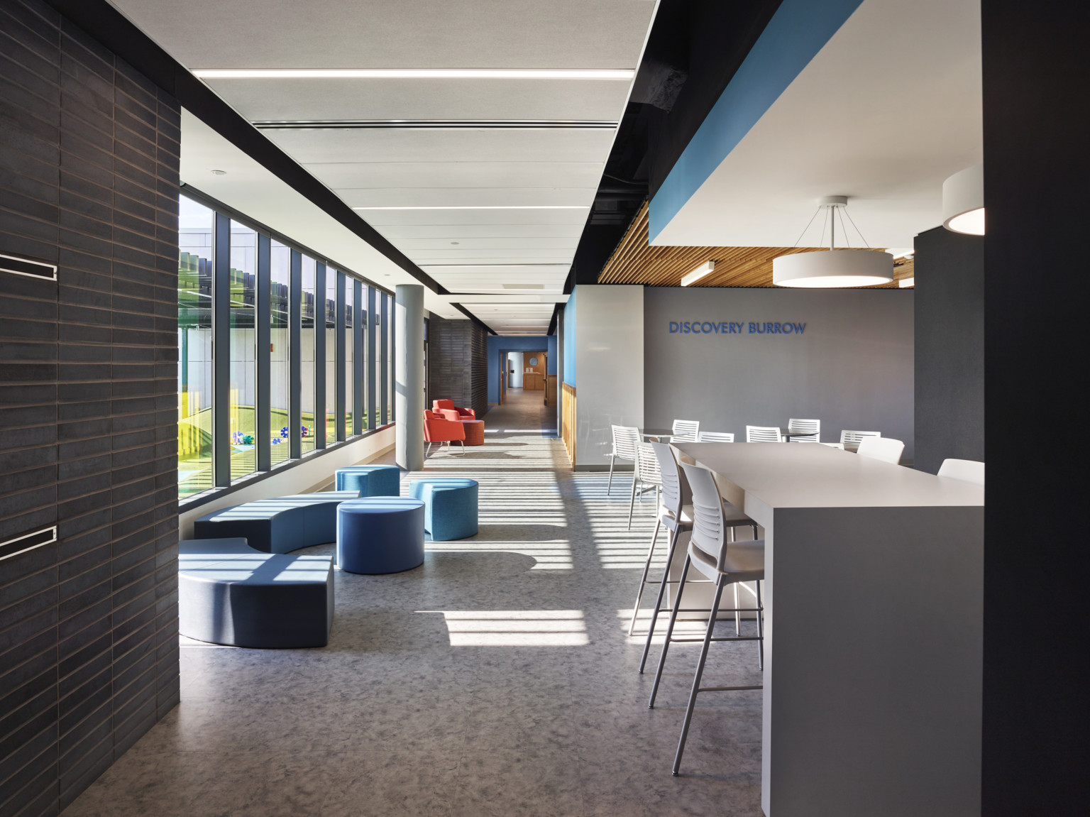 Benches and stools along windows and high top tables with stools, right, in room labeled Discovery Burrow with drop ceiling