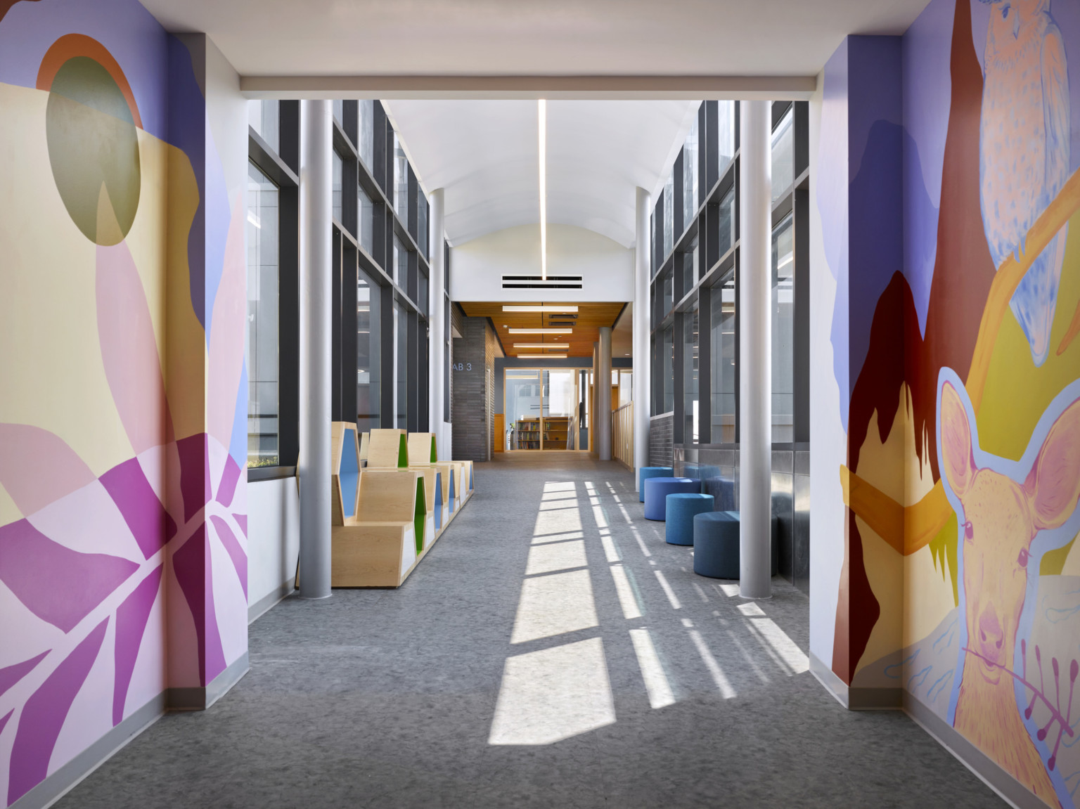 Abstract nature themed murals at entry to window lined white hallway with vaulted ceiling, wood hexagonal bench, and stools