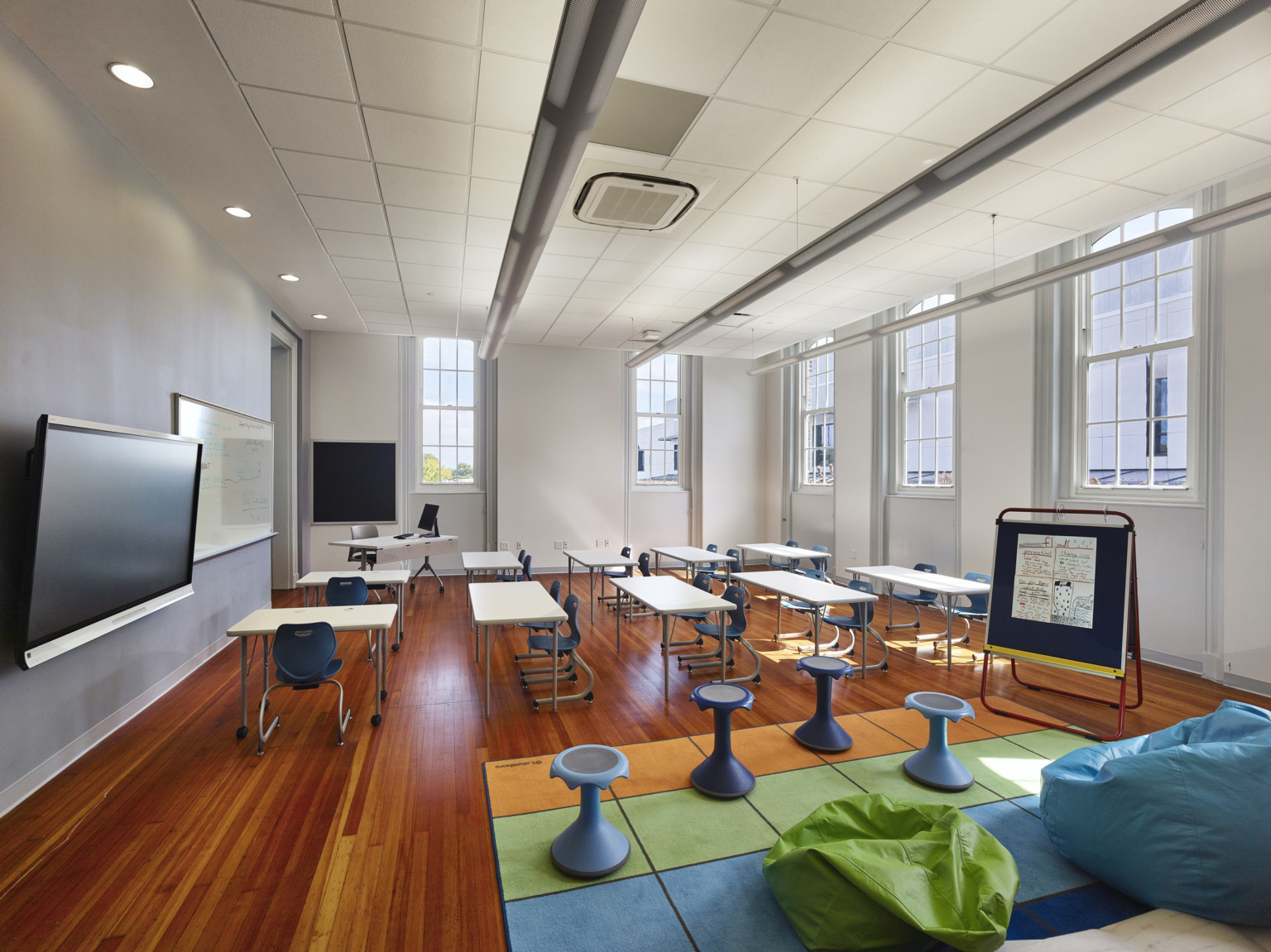 White classroom with hardwood floors and windows. Tables and chairs face screen. Nontraditional seating on colorful floor pad