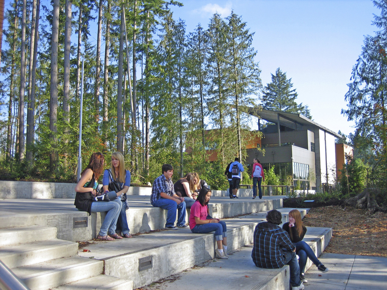 Stepped bench seating along stairs to treelined pathway. Grey building with orange accents and angled roof in background