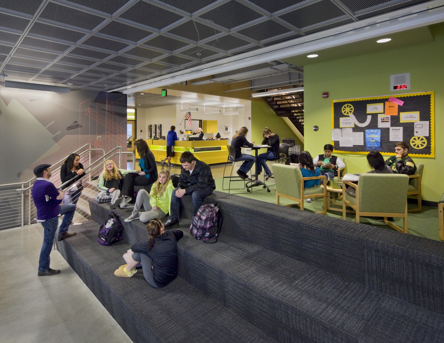 Grey carpeted step seating with stairs to left in front of green accent wall and chairs. Yellow reception desk back left