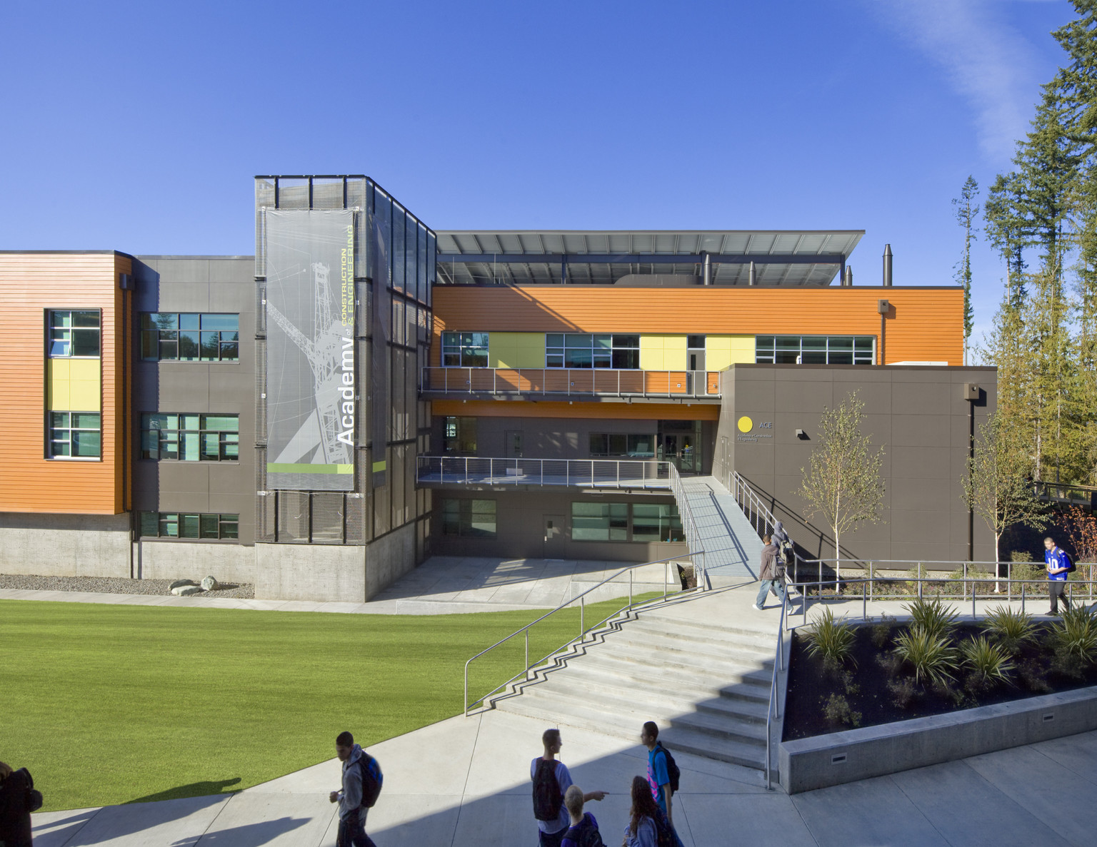 Concrete steps to L shaped ramp. Triple height glass tower extends from building, left between grey and orange facades