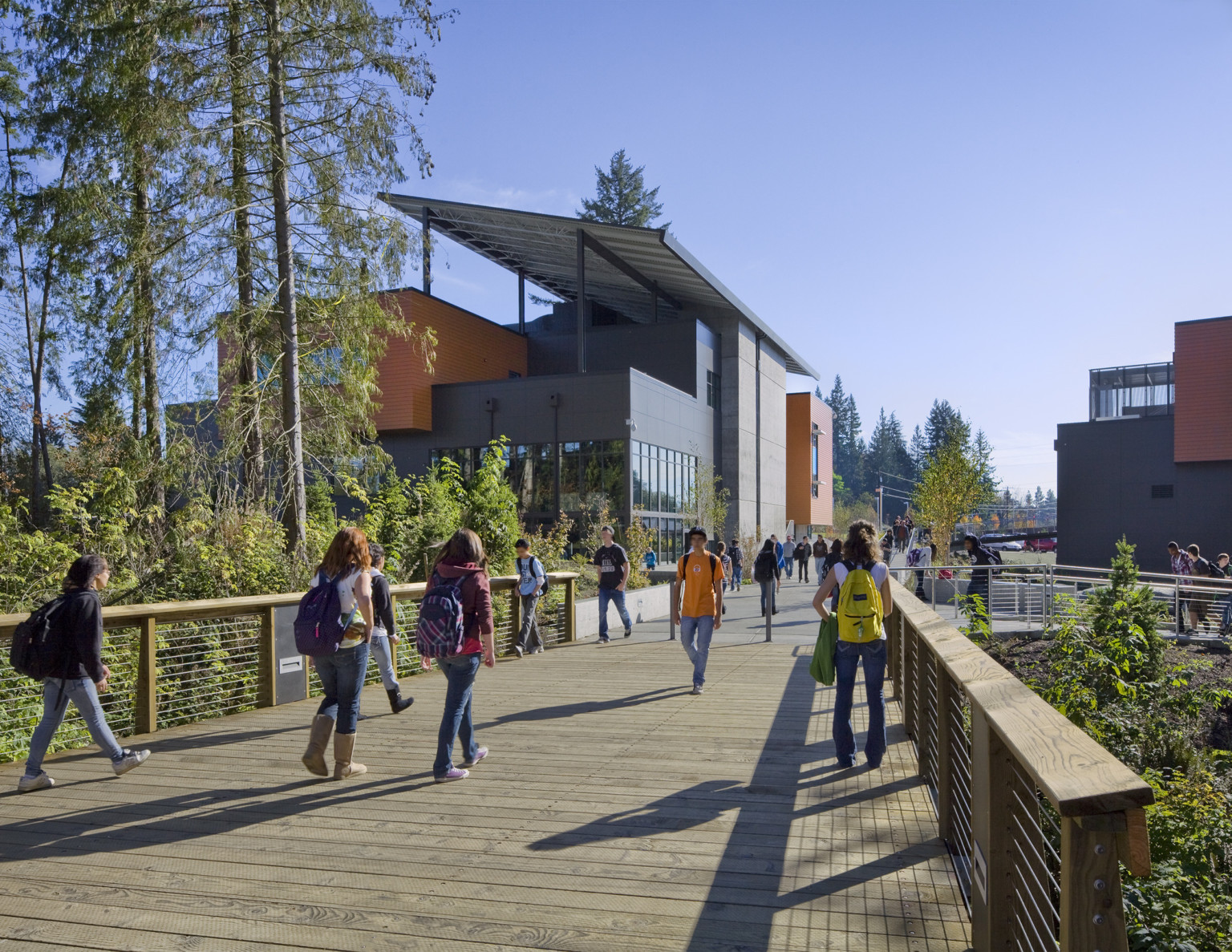 Wood bridge over trees looking to grey 3 story building with orange panel accents and large roof panel angled across top