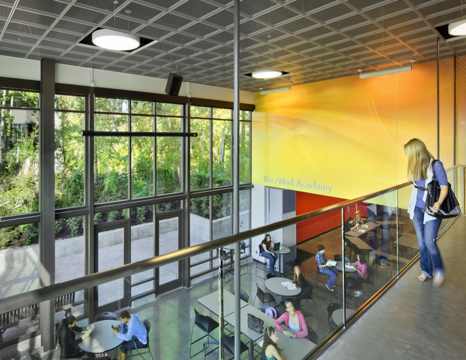 2nd floor walkway in double height atrium with windows looking out to trees. Yellow accent wall to right over seating area