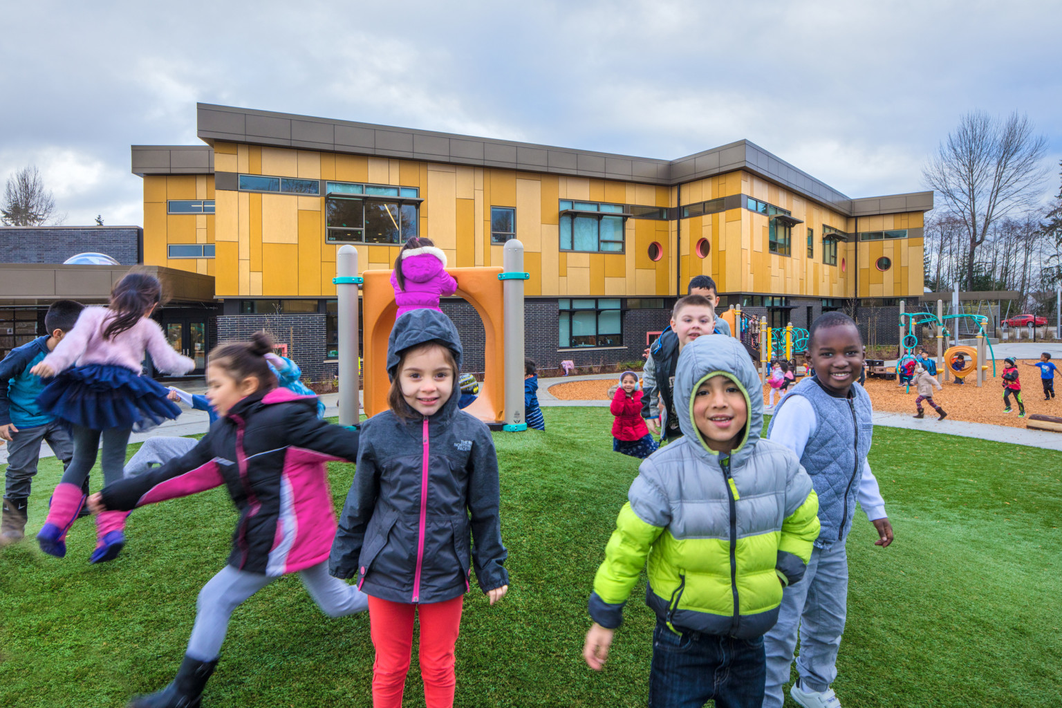 Pathfinder Kindergarten Center students on a playground in front of a building with dark bricks below a wood panel 2nd floor