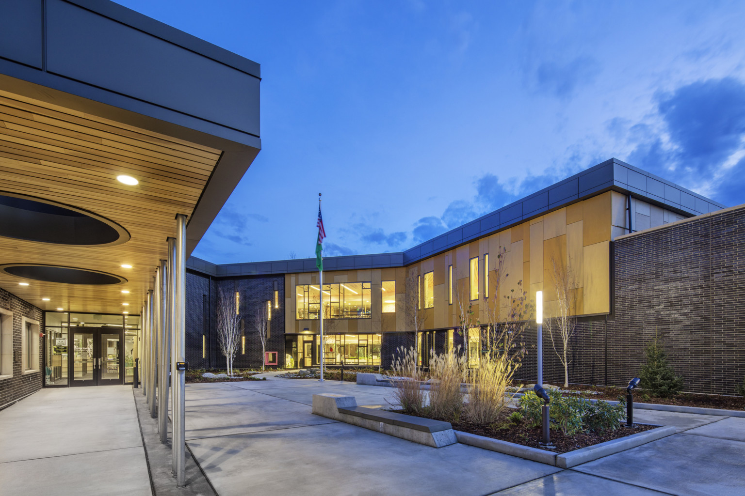 Front entry to building with patio shelter covering walkway. Right, a building wrapped courtyard with planters and flag