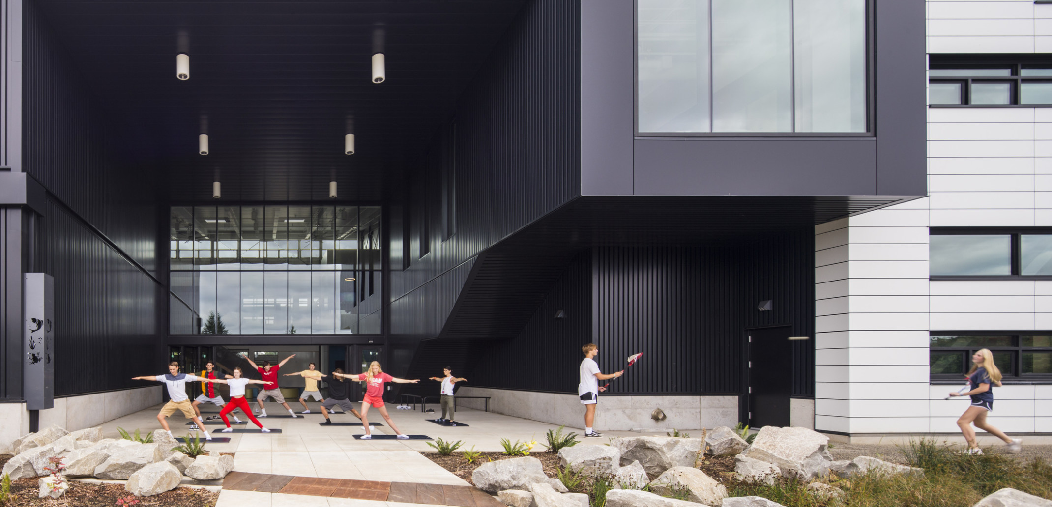 Exterior view, atrium is recess and patio is covered. Right, the building extends in a black tunnel with large window at end