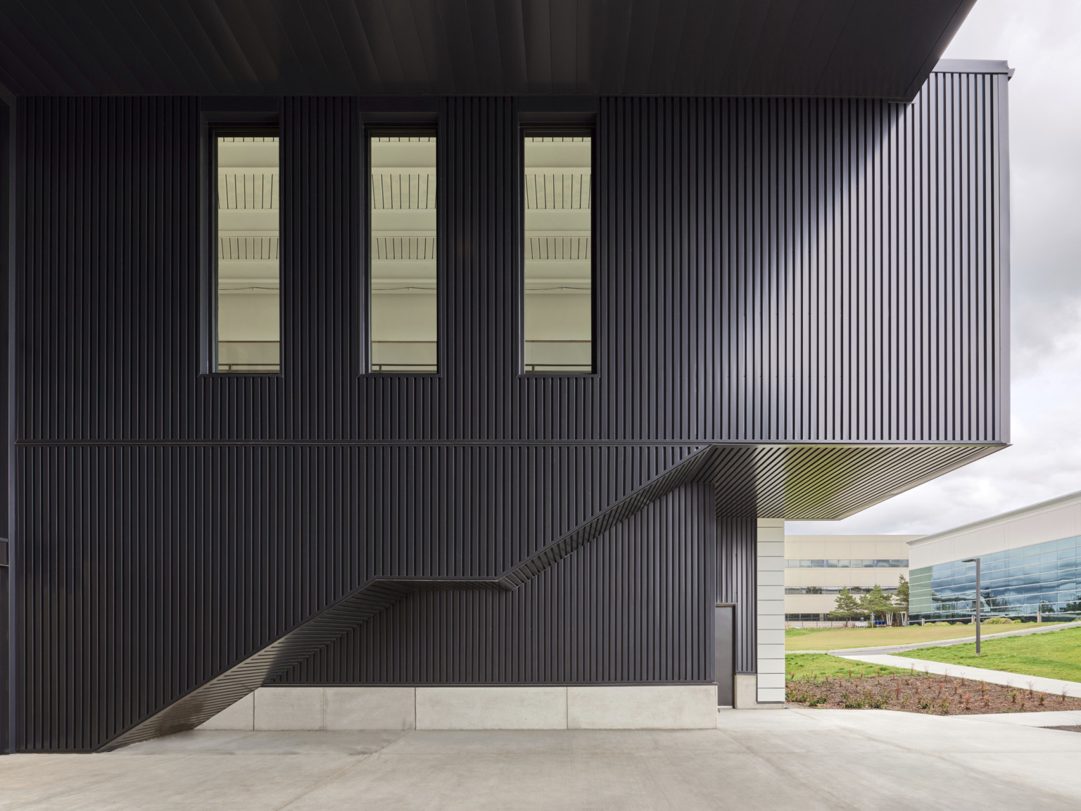 Double height black ribbed wall of covered patio with 3 long thin windows at 2nd level