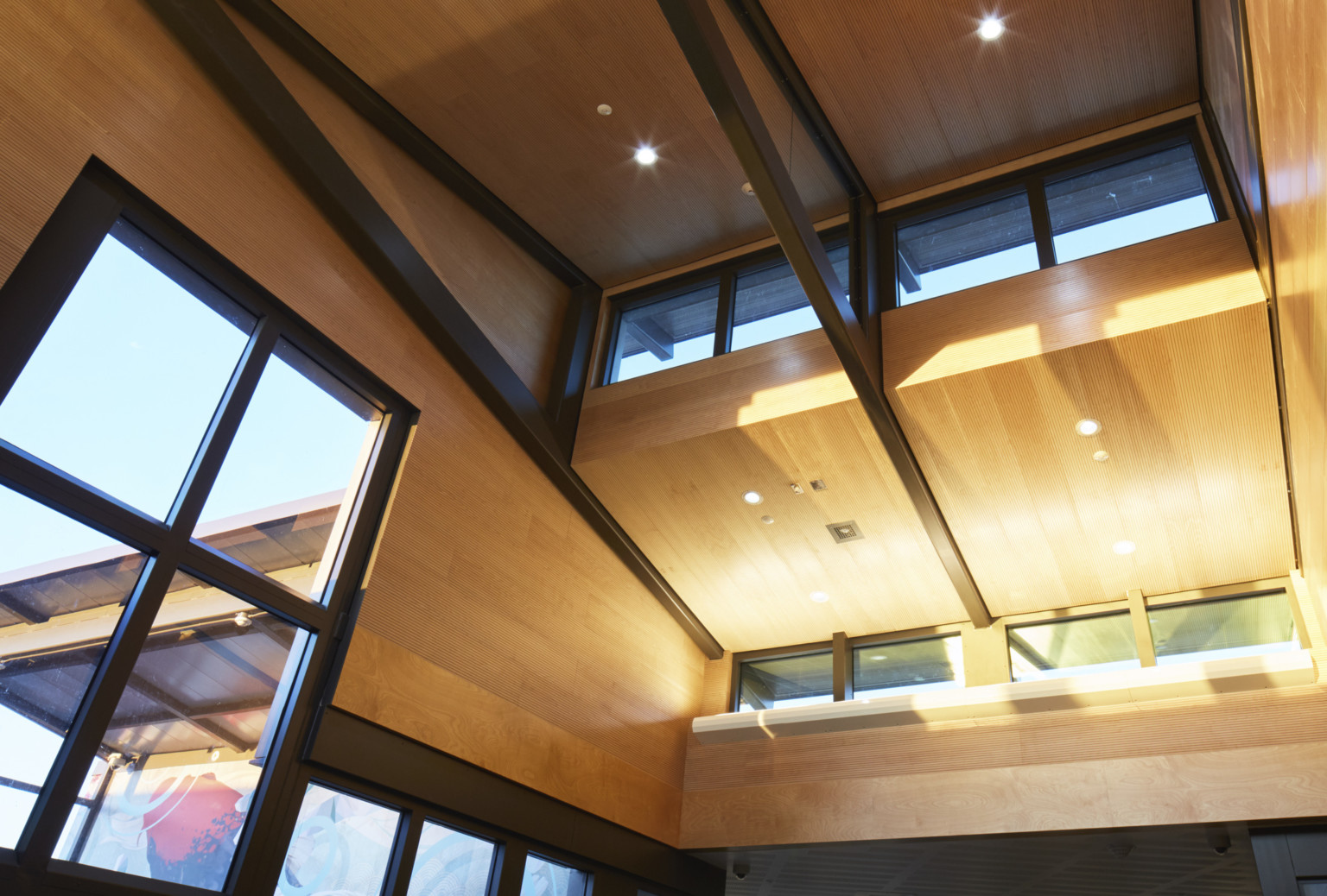 Interior view of double height wood panel hallway with large black framed windows and black trusses on drop ceiling detail