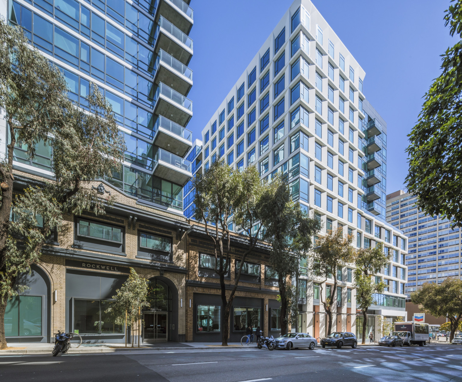 Glass building with white wrap facade and dark geometric seams. Balconies on upper levels. Connected 2 story brick building