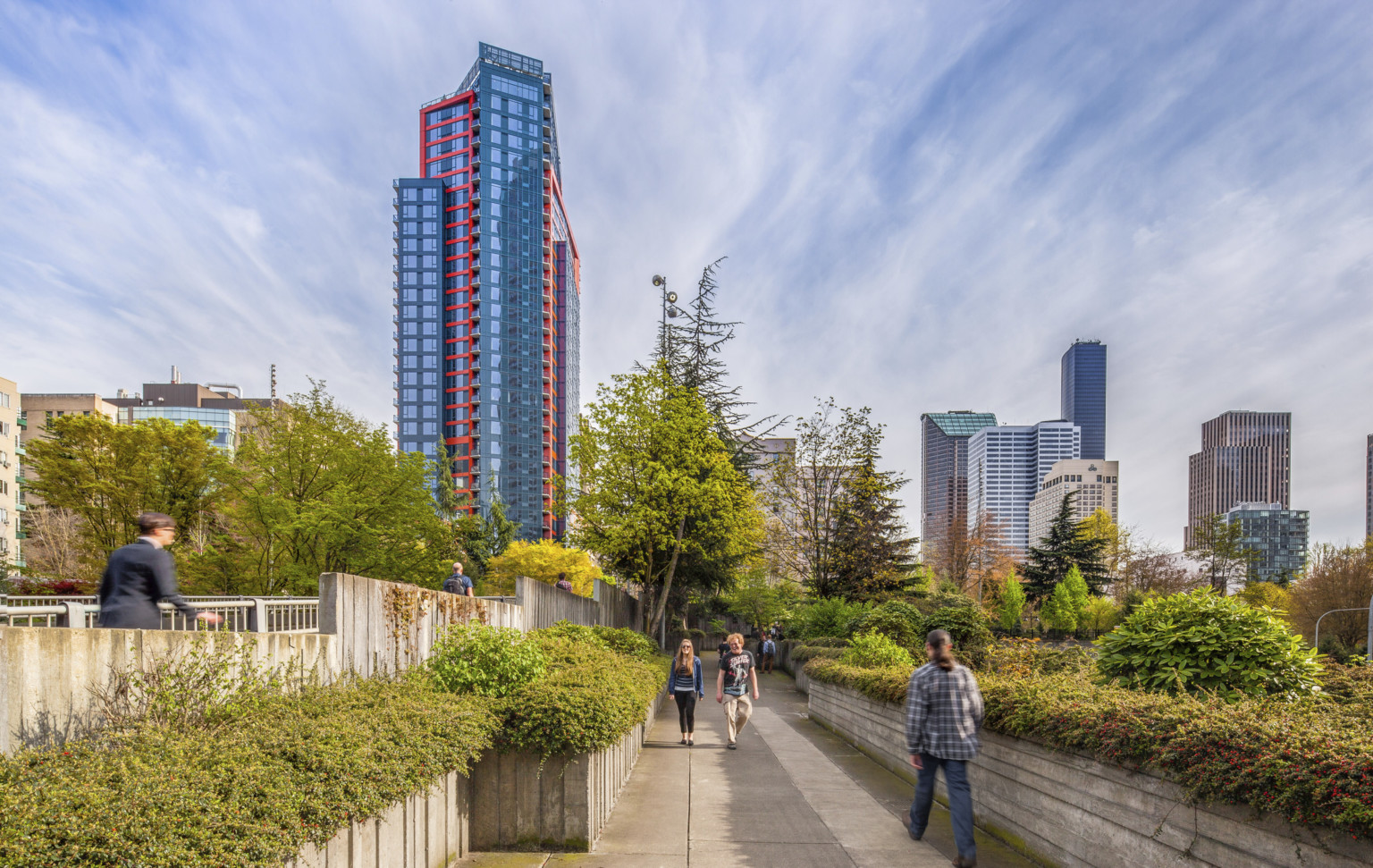 Cielo, a layered glass building with red and blue wrapping facade, behind a tree lined walkway surrounded by skyscrapers