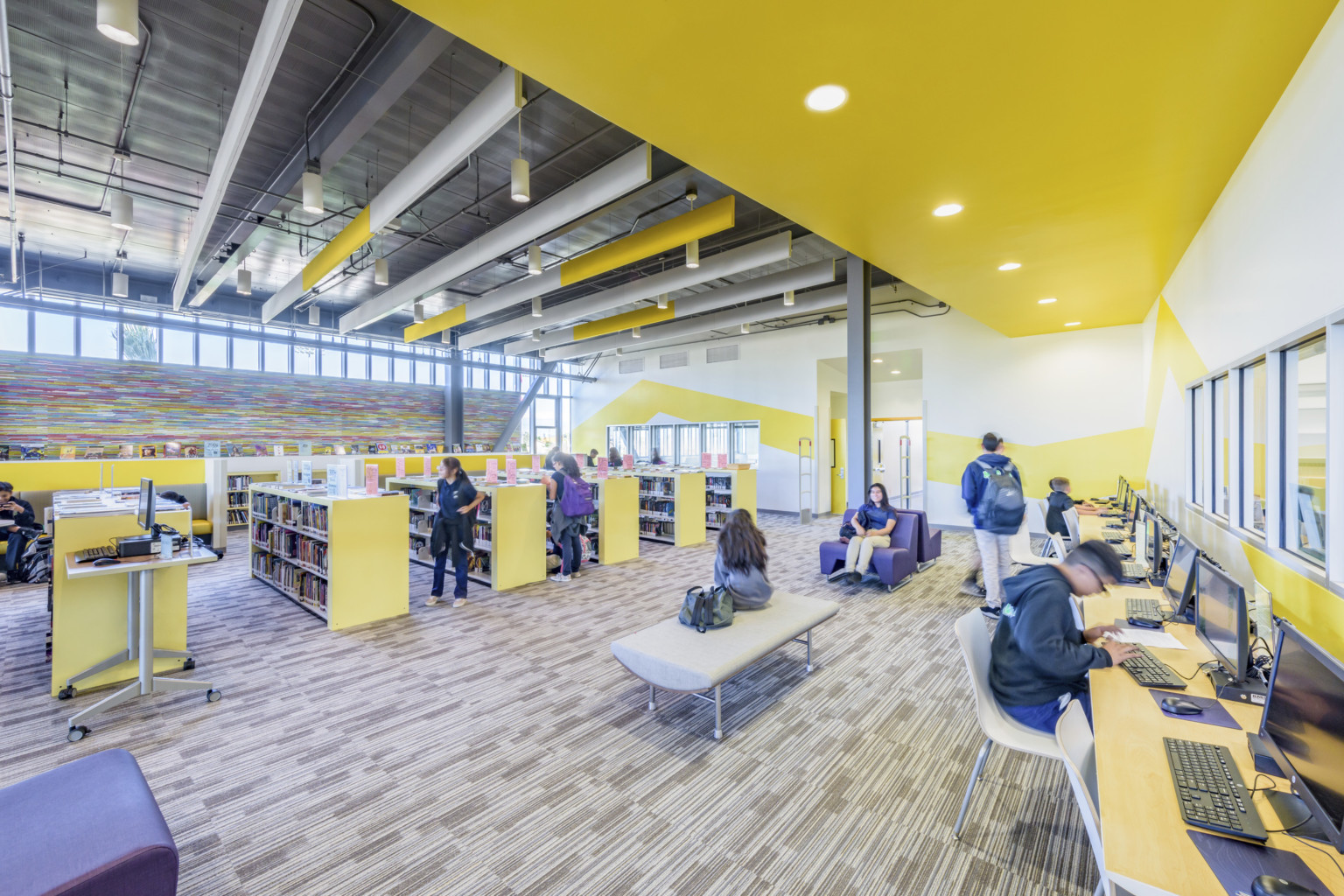 Yellow half height bookshelves in room with yellow stripes on white wall and colorful abstract pattern, left. Computers right