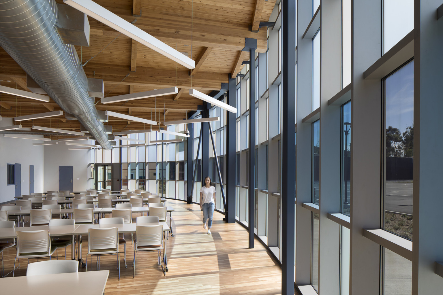 Interior view with curved glass wall, right. Left, a seating area with tables under wood ceiling at Campus Kilpatrick Youth Facility