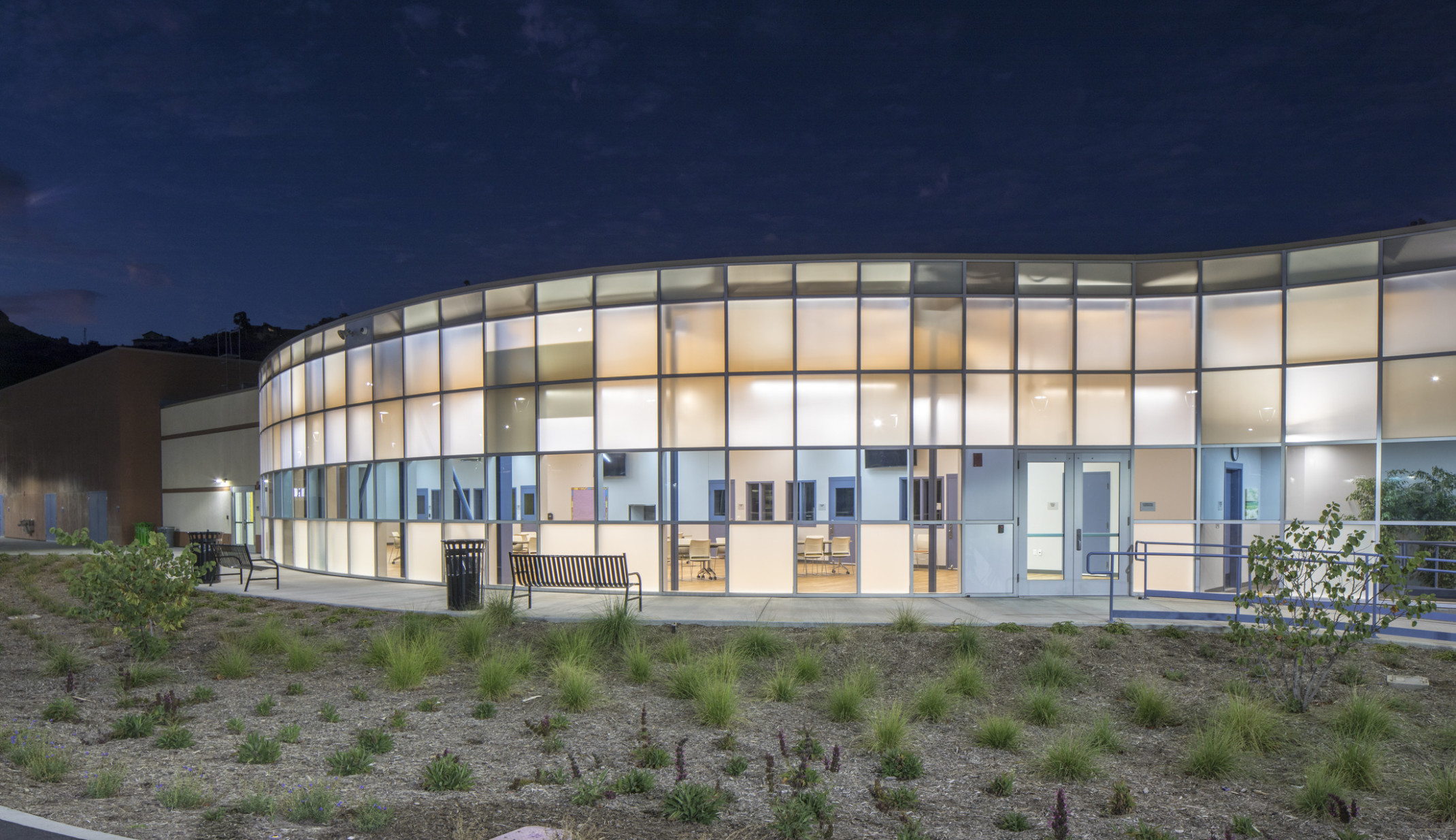 Campus Kilpatrick Youth Facility, an illuminated rounded double height paneled glass facade with blue translucent stripe Campus Kilpatrick Youth Facility, an illuminated rounded double height paneled glass facade with blue translucent stripe