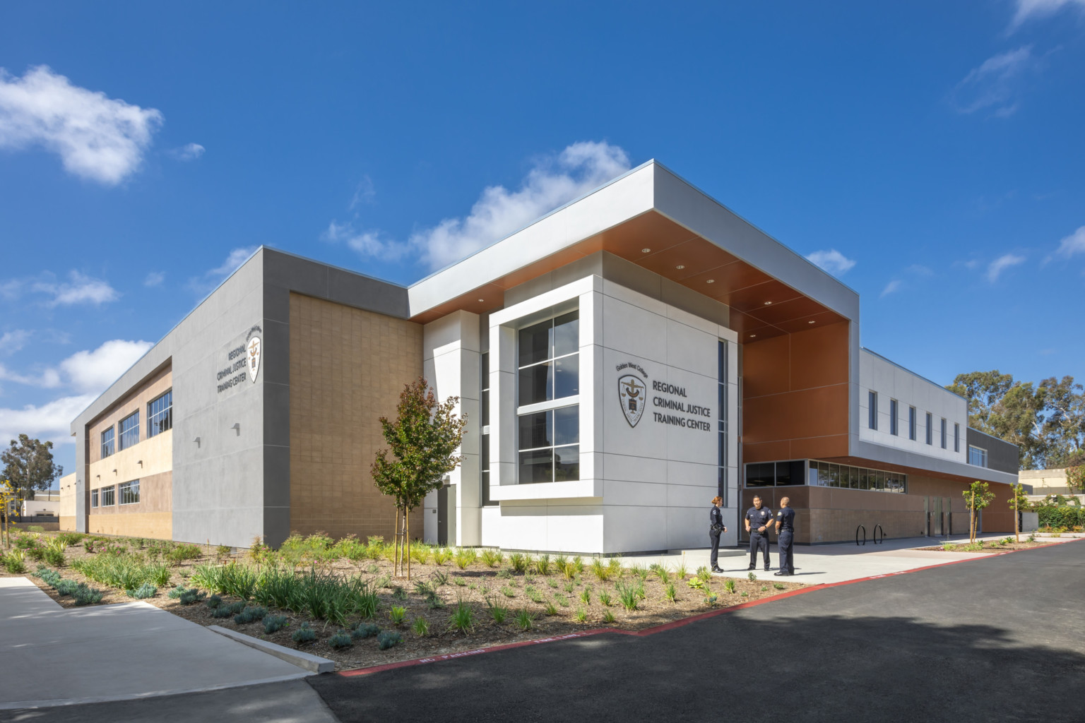 Corner exterior view of white cubic entrance with logo facing the street, shaded by a grey overhang with orange base