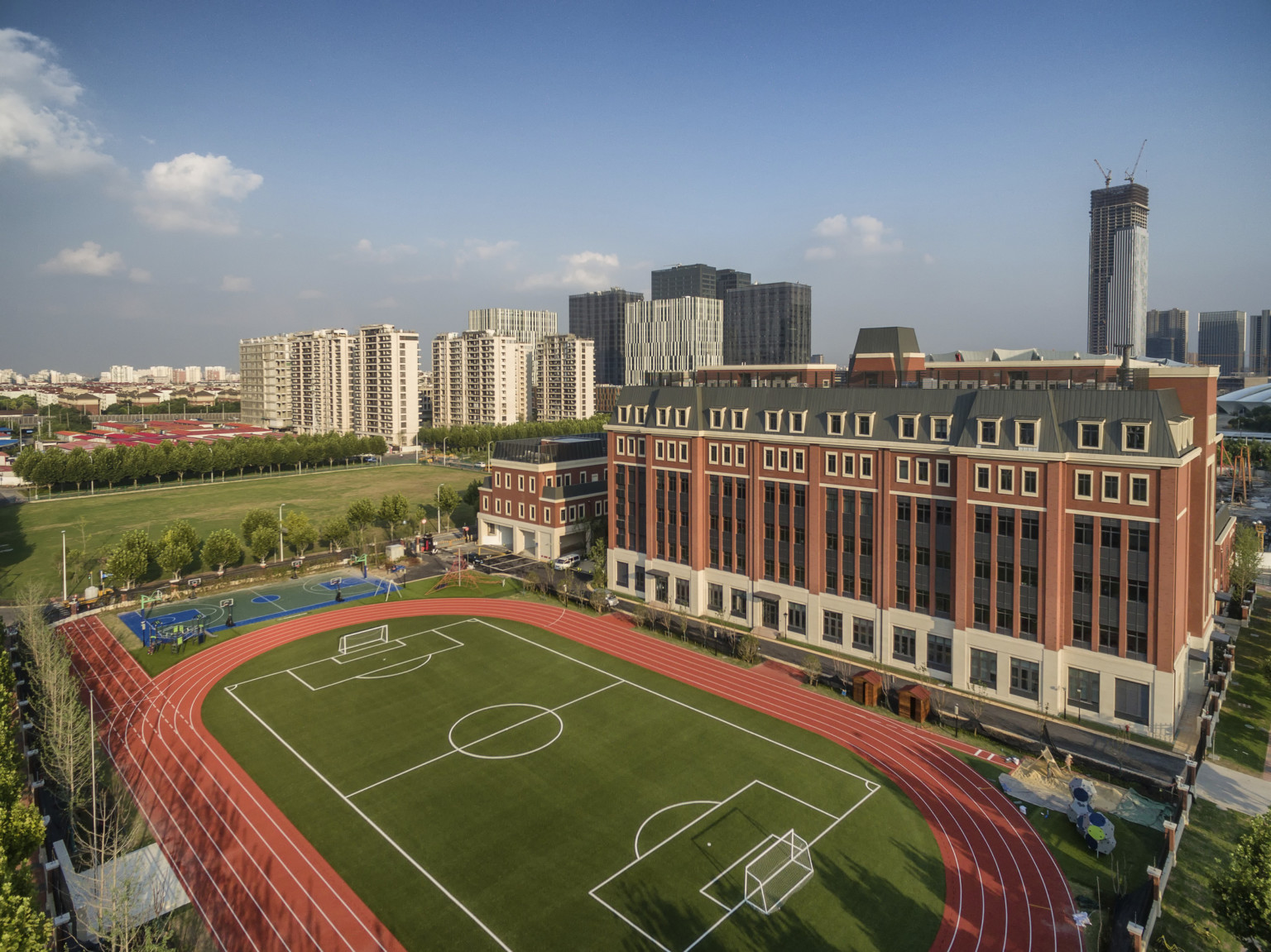 Aerial view behind school. A large 5 lane track wraps around a soccer field. Right is the school, a 6 story brick building