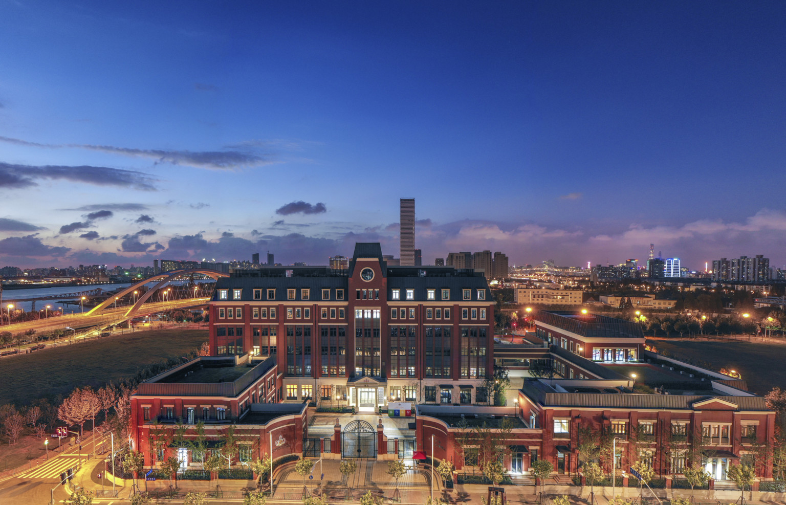 View of brick school with dark stone roof. A central tower extends above entry of the main building, illuminated at night. View of brick school with dark stone roof. A central tower extends above entry of the main building, illuminated at night.