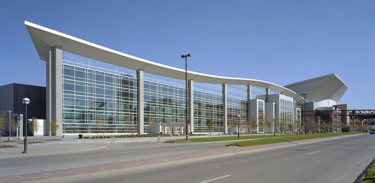 Chi Health Center Omaha has a glass facade under curving white roof with overhang. Black exposed beam walkway over the street