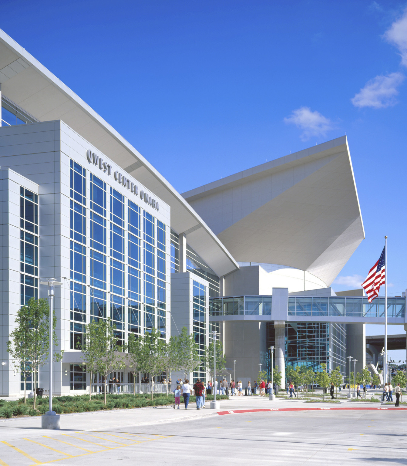 Glass skywalk connects to silver paneled building with large windows. Building labeled Qwest Center Omaha. Angled roof behind