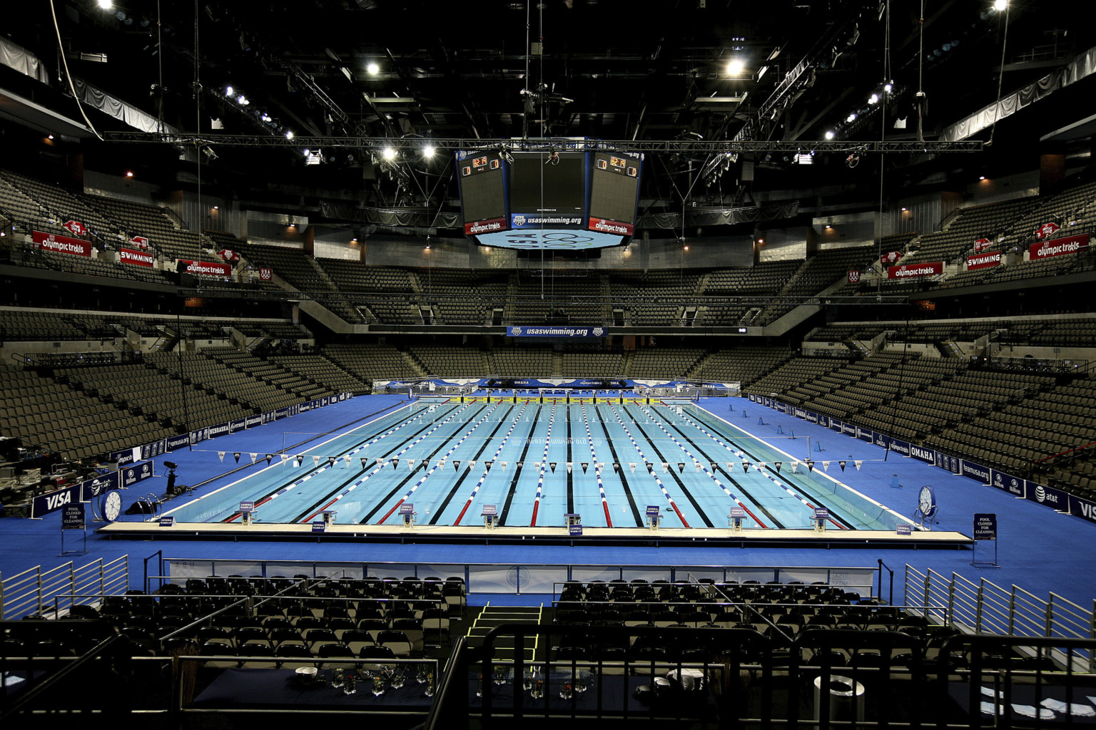 A temporary Olympic sized swimming pool set up within the empty arena. Jumbotron above has Olympic rings on bottom