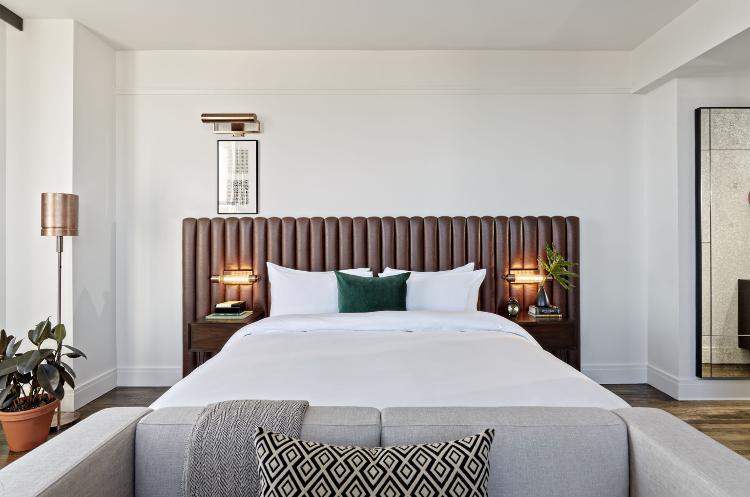 White bed with green throw pillow against a brown ribbed headboard. Side tables and lamps on each side, grey couch at base