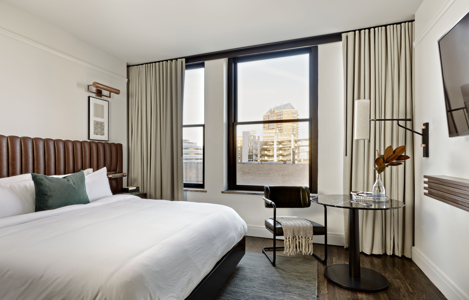 White room with bed with brown headboard, round black table and black cesca chair next to window with view of tall buildings.