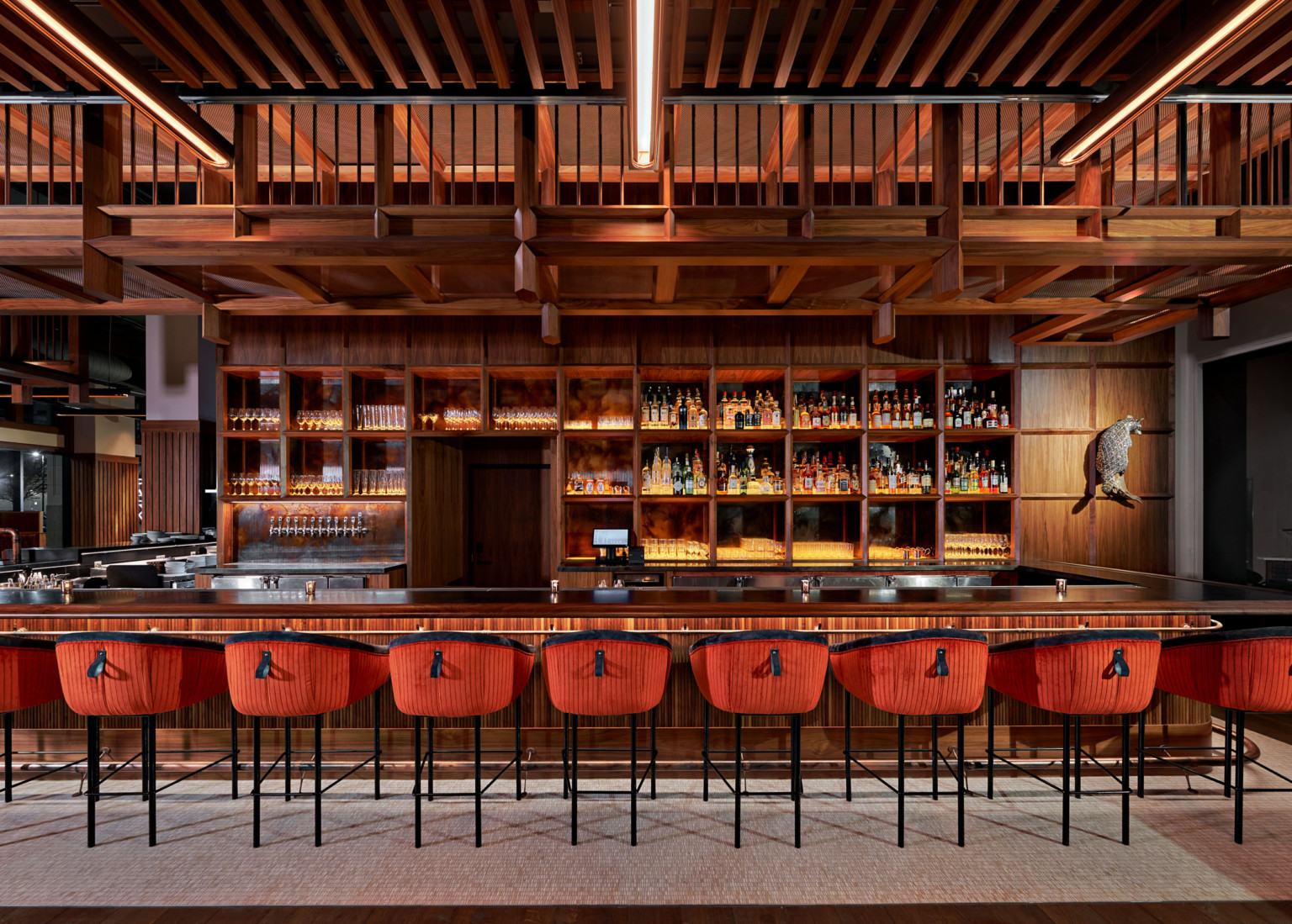 Large wood bar with built in shelves along the back. Wood overhang over back of bar, and stools with red and black upholstery